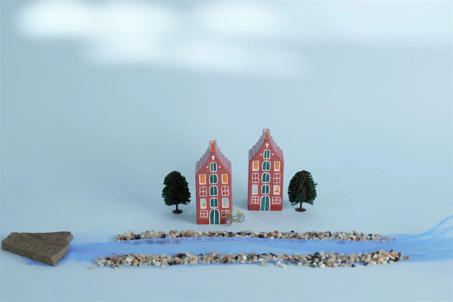 Number 02 of a Collectible Series | The Amsterdam House features two miniature Dutch-style houses with classic Amsterdam architecture and trees, set on a light background with pebbles, blue fabric, and a flat stone in the foreground.