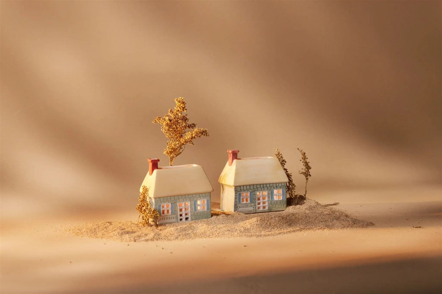 Number 04 of a Collectible Series | The English Cottage: This charming ceramic house with lit windows sits amid dried plants on sandy ground against a beige background—ideal as unique decor or a tea light holder for your space.