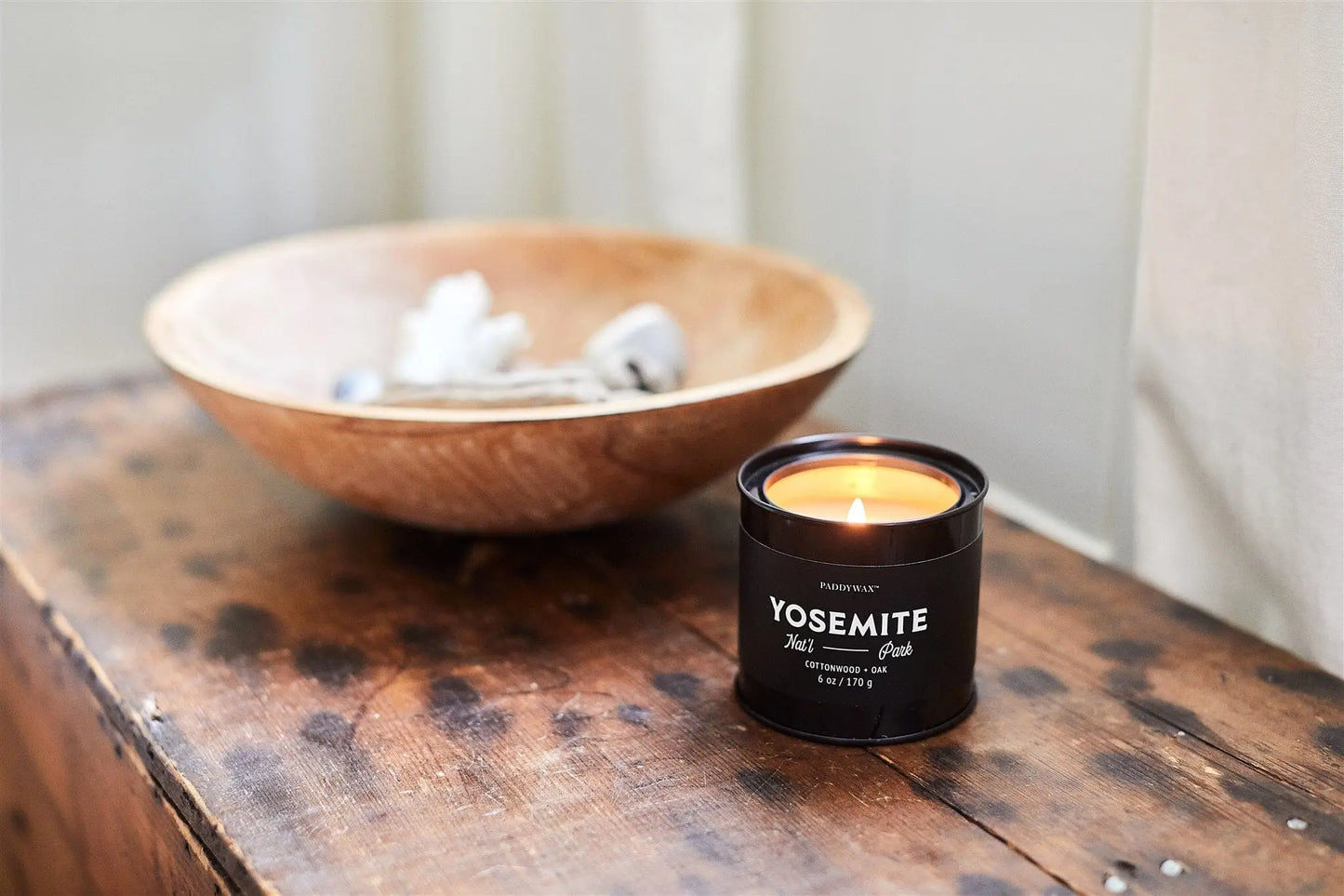 A lit Parks 6oz Tin - Yosemite candle sits on a wooden surface beside a bowl of small white objects—perfect for any outdoor enthusiast.