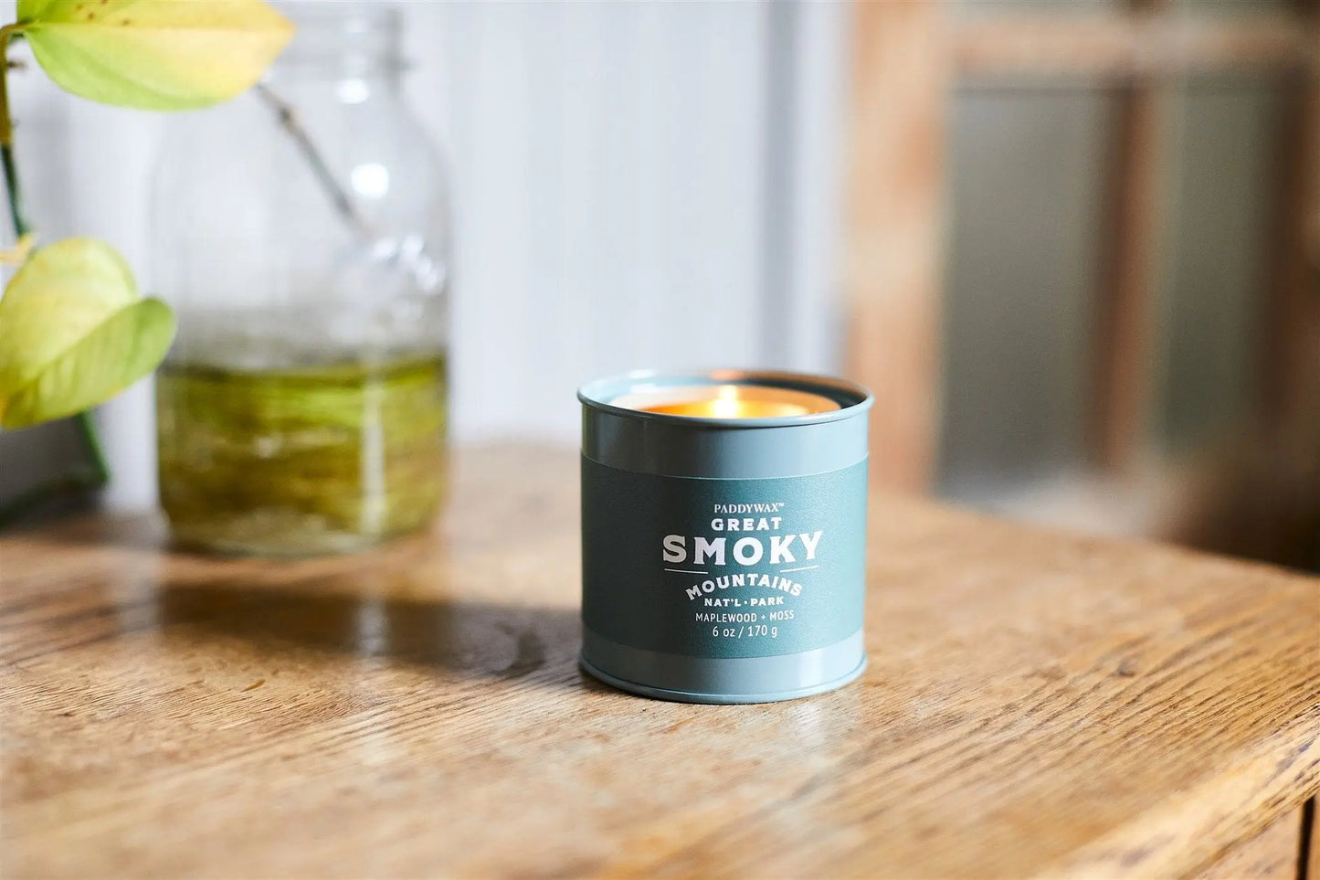 A lit Parks 6oz Tin - Great Smoky Mountains candle sits on a wooden table beside a glass jar with green liquid and a plant cutting, capturing the tranquil essence of the renowned US National Park.