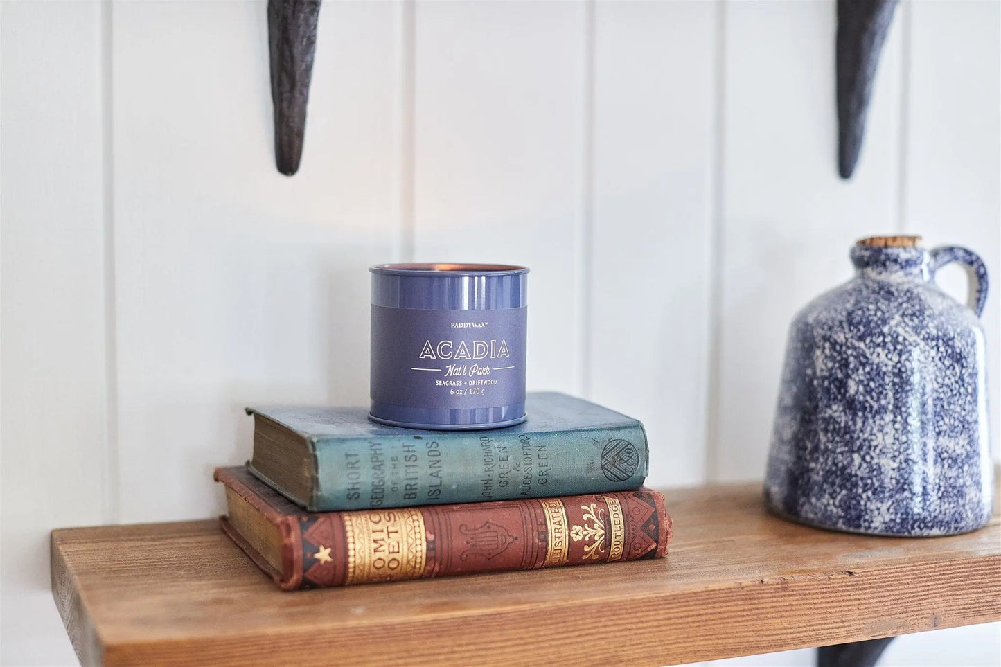 A lit Parks 6oz Tin - Acadia candle from the Parks Collection rests on two vintage books atop a wooden shelf beside a blue and white ceramic jug.