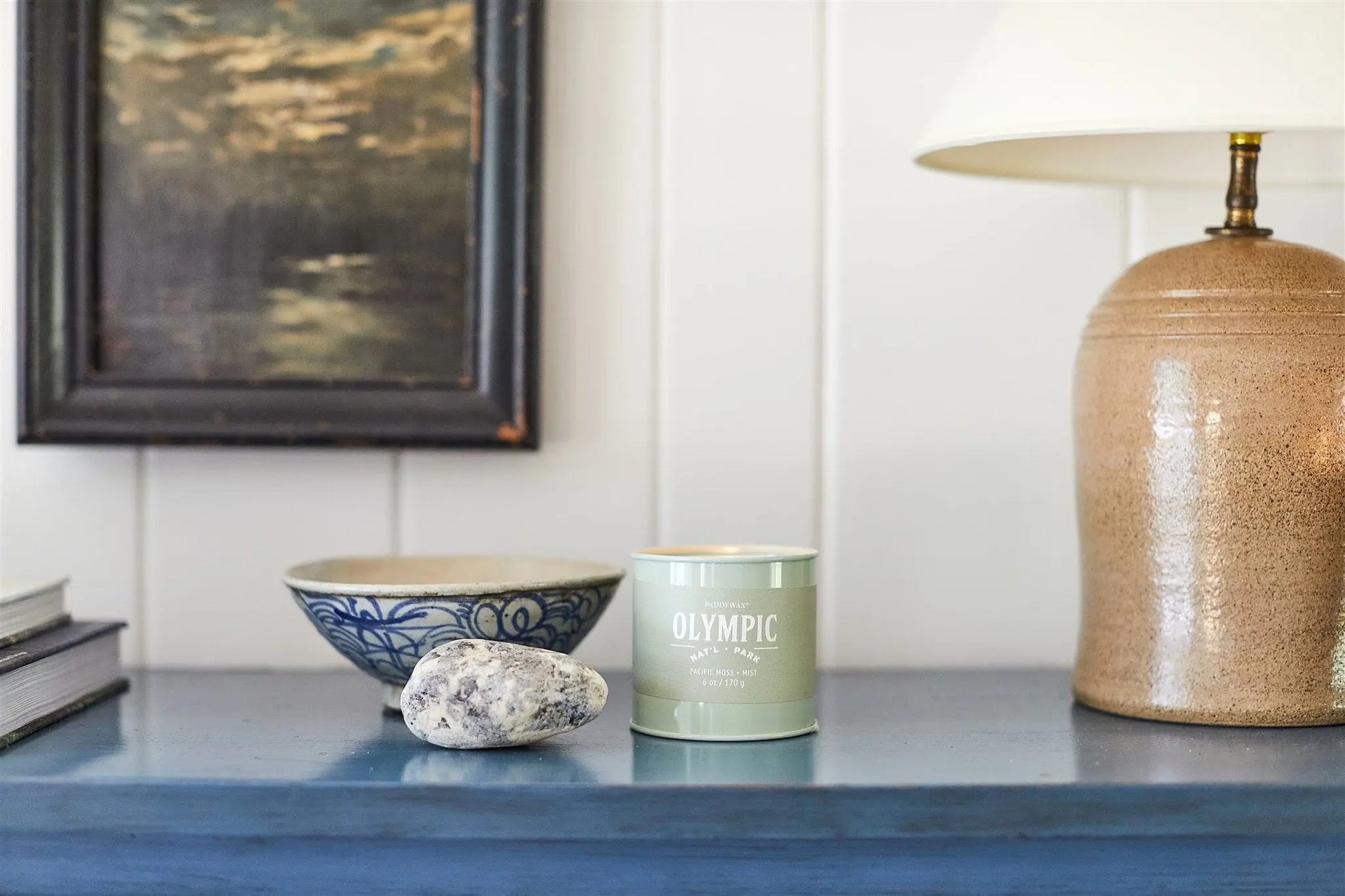 A Parks 6oz Tin - Olympic candle sits on a blue table near a speckled stone, ceramic bowl, lamp, and framed painting on a white wall—ideal for any outdoor enthusiast.