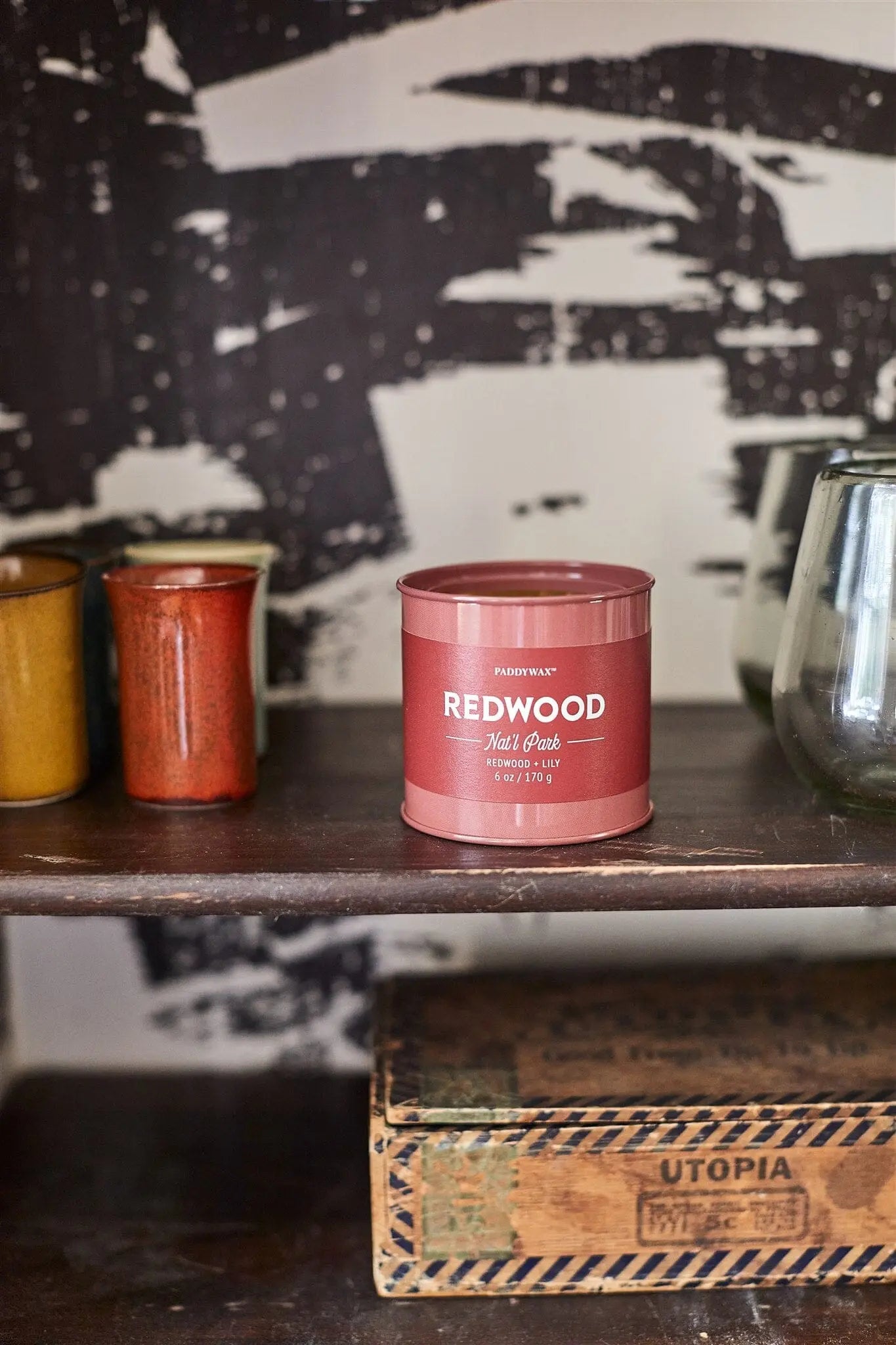 A Parks 6oz Tin - Redwood from the Parks Collection sits on a wooden shelf beside ceramic cups and a clear glass, above a vintage box labeled Utopia.