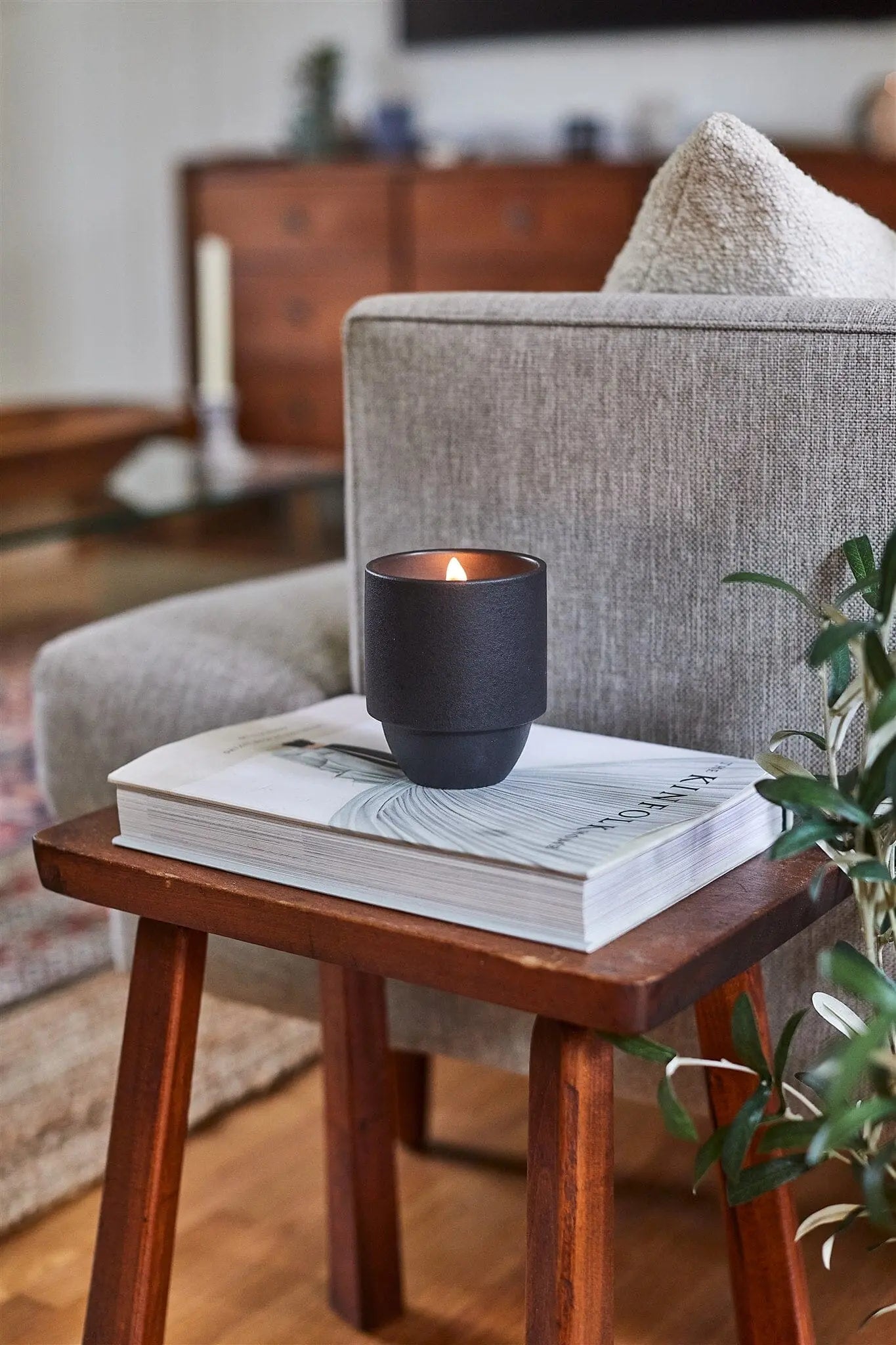 A lit Parks 11 oz Candle - Yosemite rests on a closed book on a wooden side table beside a gray armchair in a cozy living room.