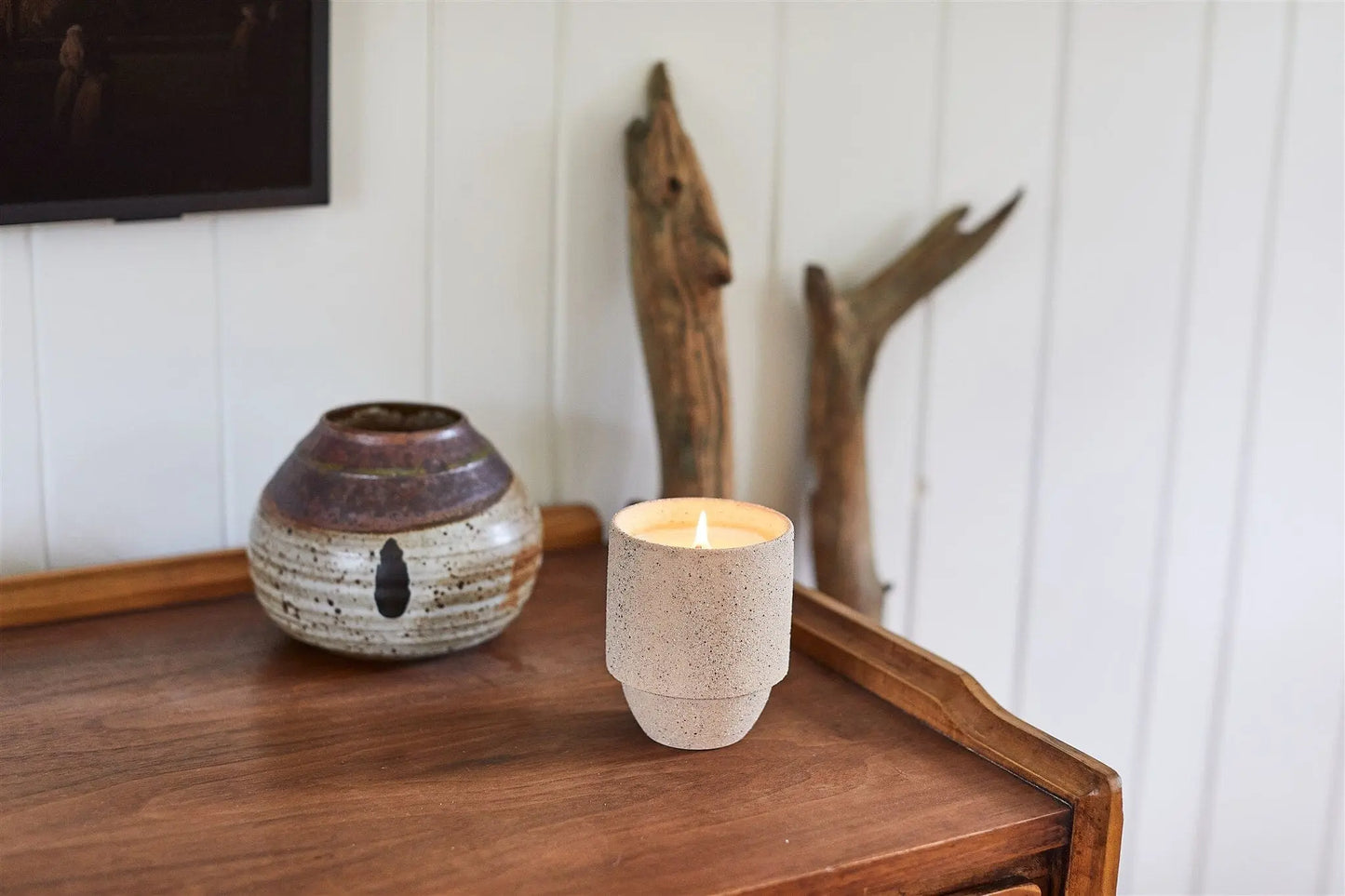The Parks 11 oz Candle - Yellowstone, in a speckled holder with crackling wood wicks, sits on a wooden surface near a ceramic pot and driftwood, capturing the rustic charm of national parks against a white paneled wall.