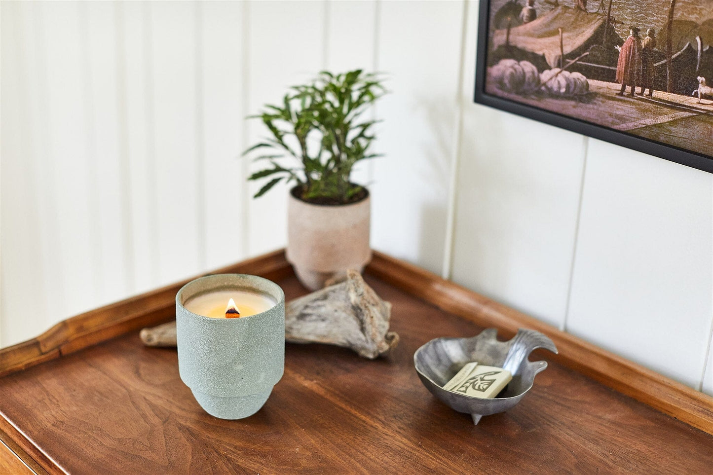 A Parks 11 oz Candle - Olympic with crackling wood wicks is lit on a wooden surface, along with a small potted plant, driftwood, and a decorative dish of matches beneath a framed wall picture.