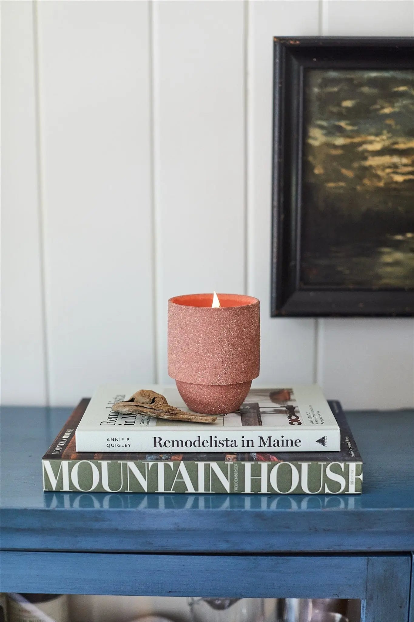 A lit Parks 11 oz. Candle - Redwoods with crackling wood wicks sits on two stacked books, Remodelista in Maine and Mountain House, on a blue table by a white paneled wall and framed painting.