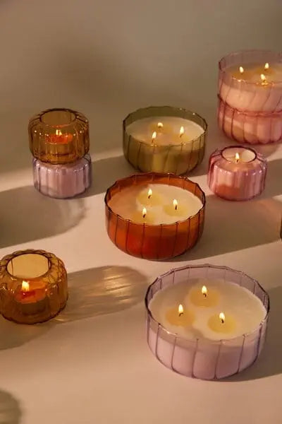 Several lit Ripple Candle - Salted Iris in ribbed, colorful glass vessels of various sizes are arranged on a flat surface, casting soft shadows.