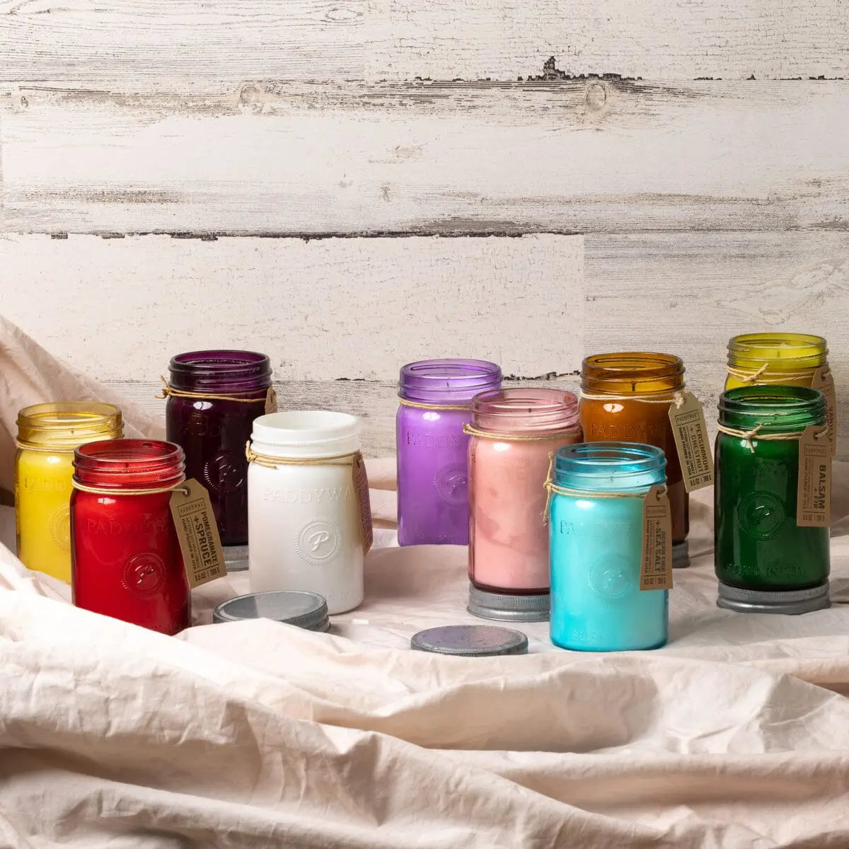 Nine Relish 9.5 oz Candles in Salted Grapefruit, each in a colored jar with metal lid and tag, are arranged on a white cloth before a rustic wooden wall—ideal for fragrance display or as charming décor accents.