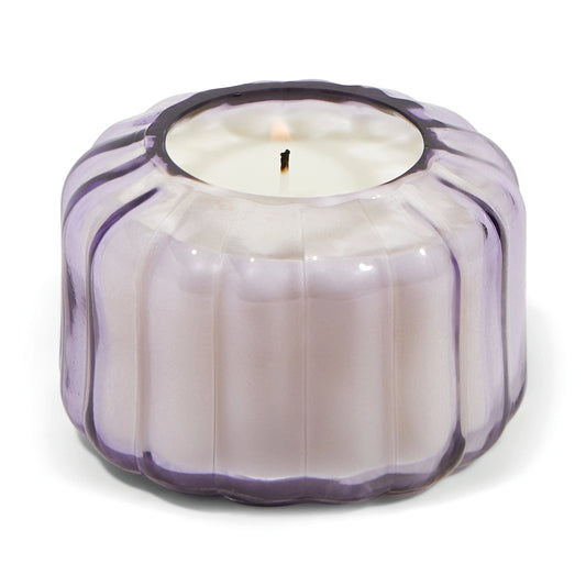The Ripple Candle - Salted Iris glows in a round, ribbed, translucent lavender glass vessel with a lit wick.