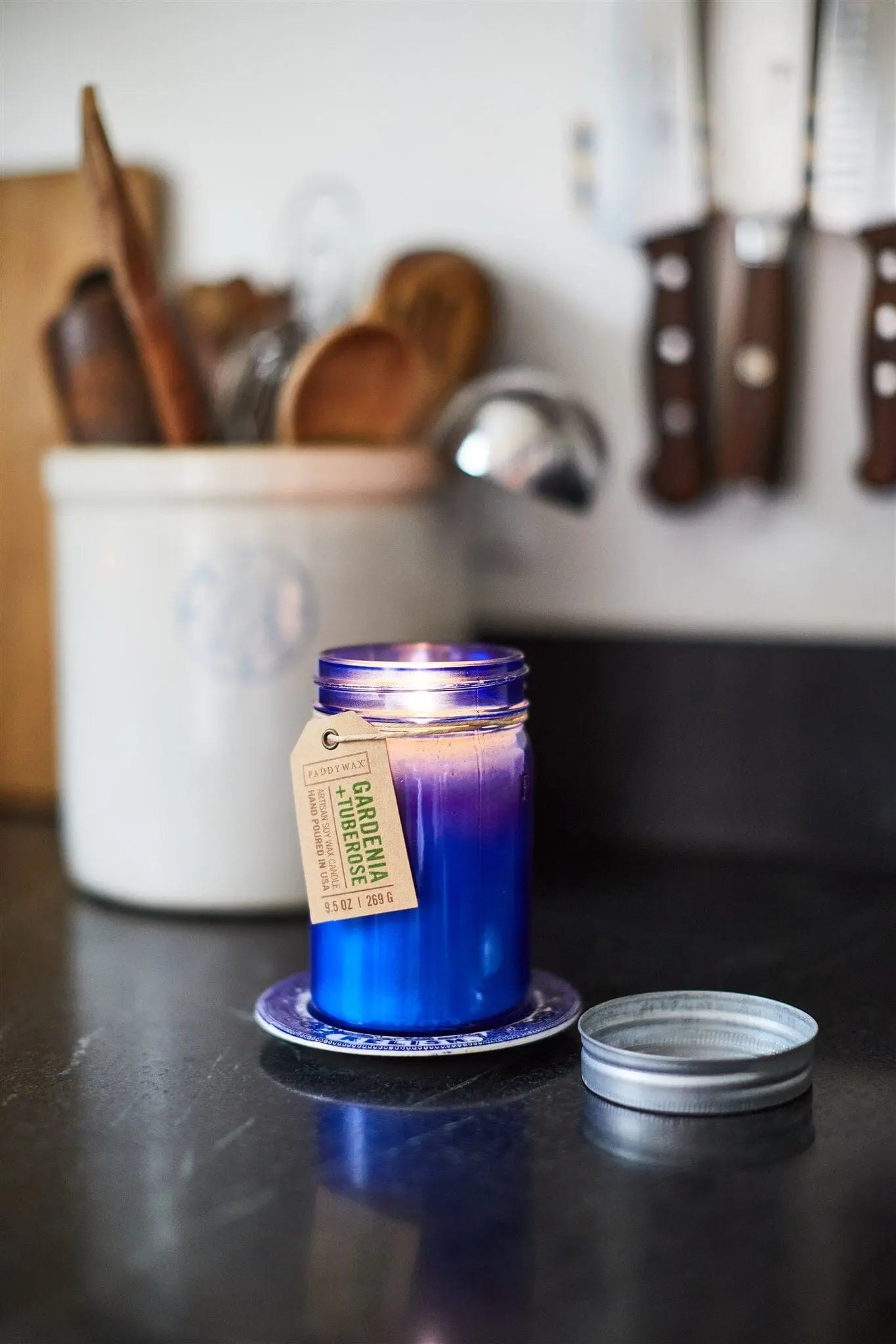 A Relish 9.5 oz Candle - Gardenia & Tuberose in a glass jar sits on a small plate on the kitchen counter, filling the room with its scent, utensils behind it and the lid placed aside.