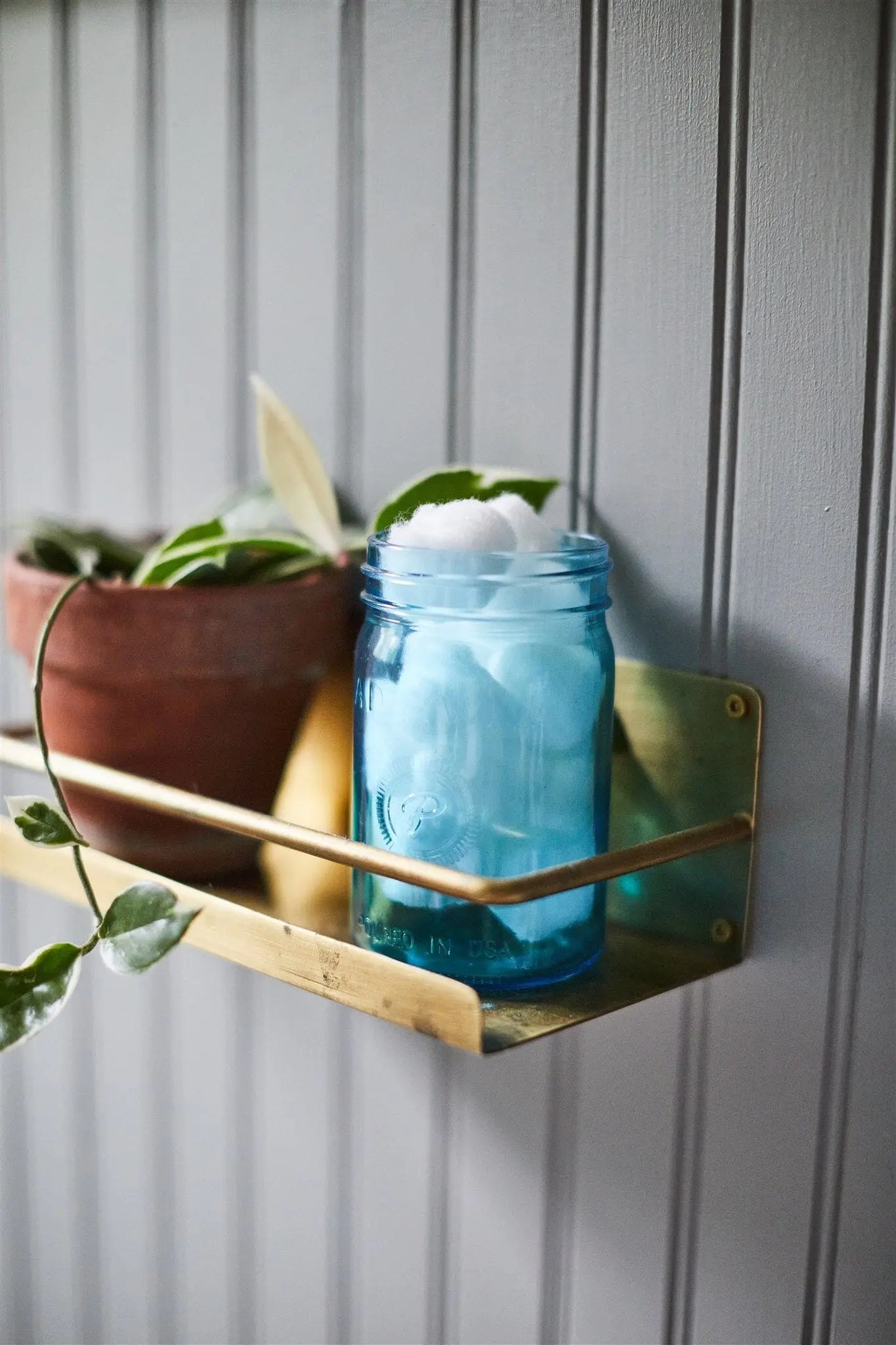 A Relish 9.5 oz Candle - Ocean Tide & Sea Salt and a potted plant sit on a brass shelf mounted to a gray paneled wall, bringing fragrance and charm to the space.