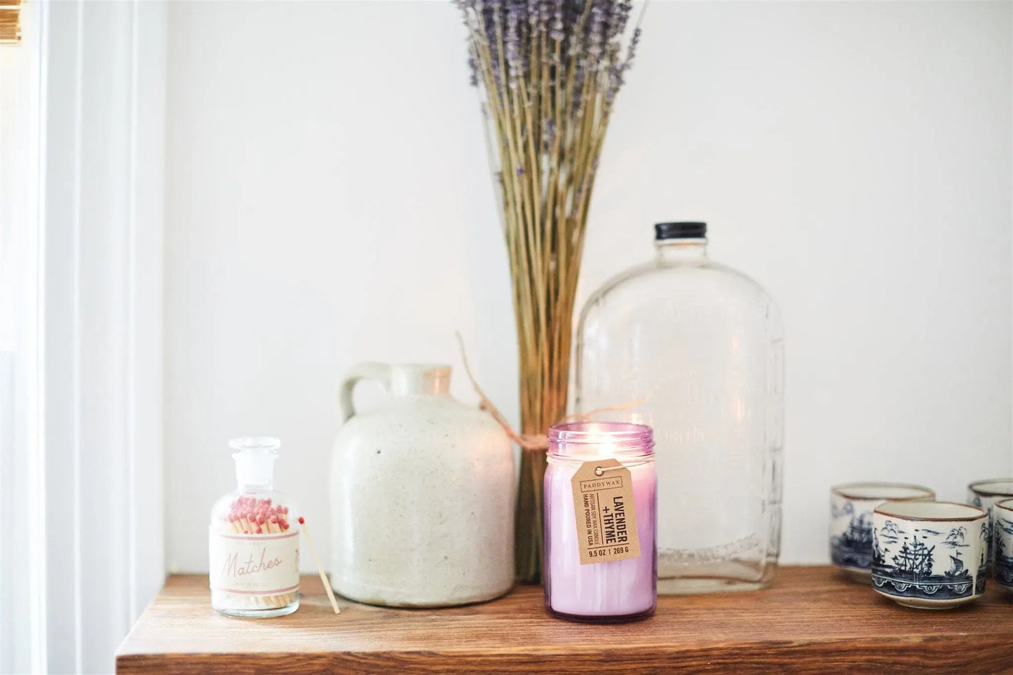 A lit Relish 9.5 oz Candle - Lavender + Thyme sits on a wooden shelf with jugs, a storage jar, dried lavender, and patterned teacups, creating a cozy atmosphere filled with gentle fragrance.
