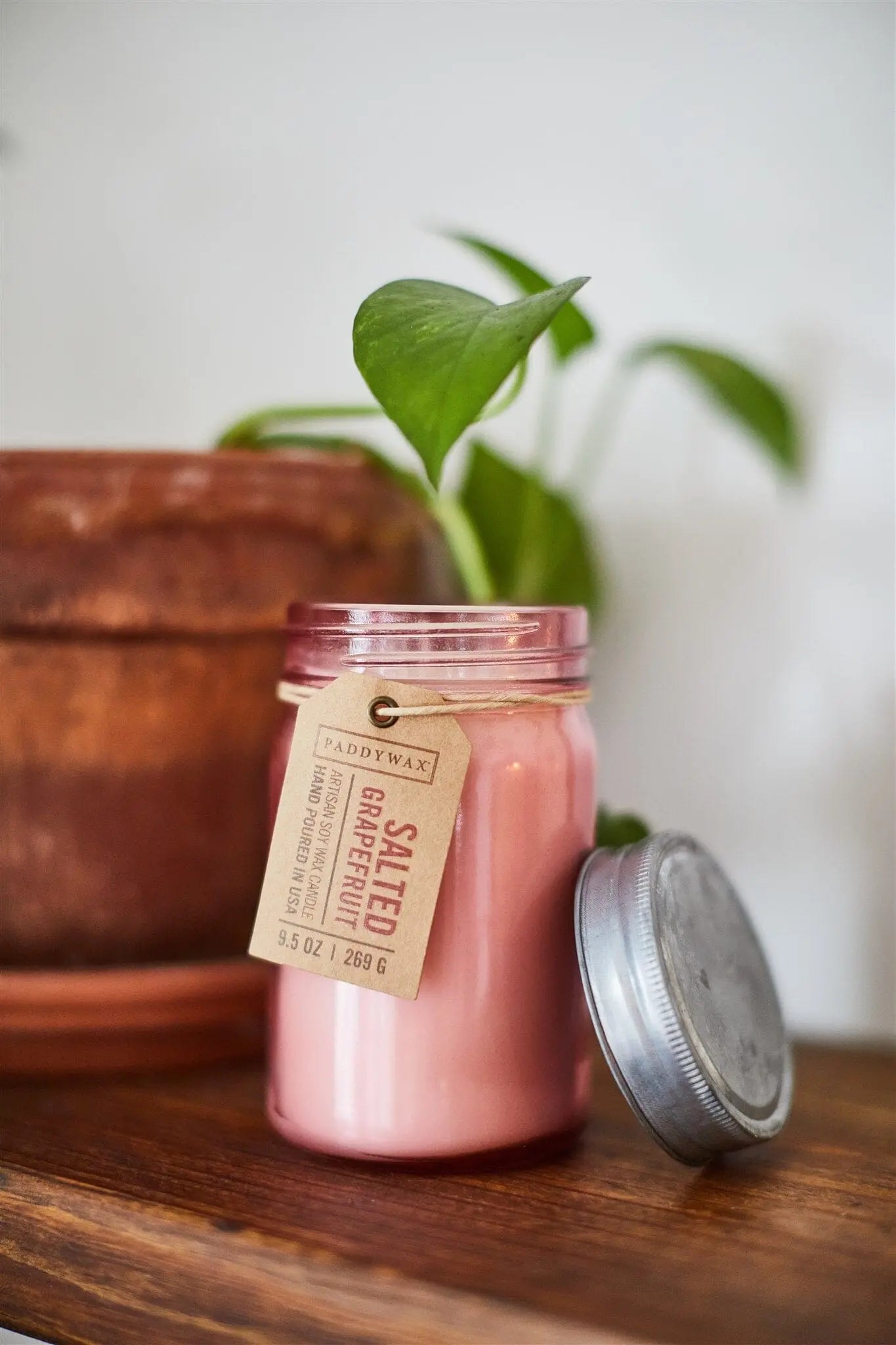 A Relish 9.5 oz Candle - Salted Grapefruit with a metal lid sits open on a wooden surface beside a brown plant pot and green leafy plant, filling the space with its refreshing, citrus-inspired fragrance.