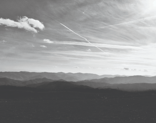 Black and white photo of distant mountain ranges under a sky with wispy clouds and several visible airplane contrails.