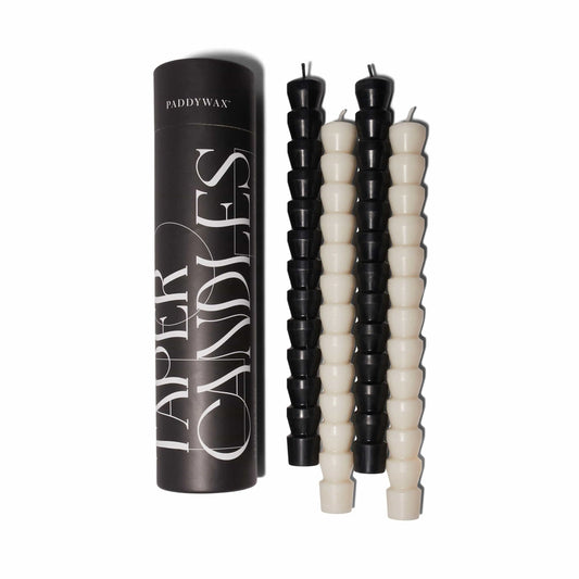 The Black & White Tube Tapers - Set of 4, featuring two black and two white ribbed candles, are displayed next to a black cylindrical box labeled Taper Candles by Paddywax on a bright white background.