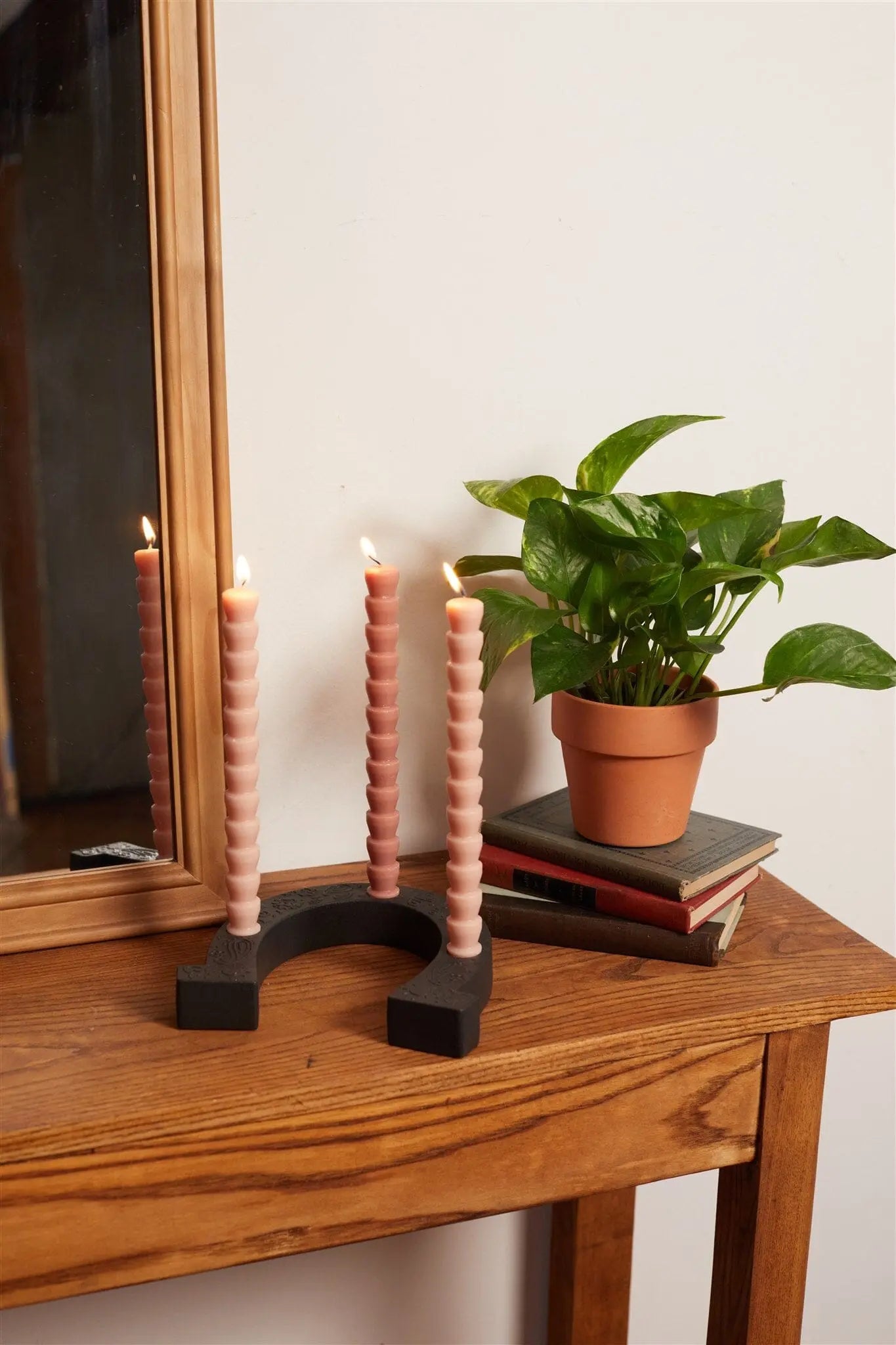 A wooden table displays a potted plant on stacked books and the Nashville Black Ceramic Horseshoe Taper Holder w/ Hangtag, topped with four lit pink spiral candles, beside a framed mirror—an inviting home decor accent.