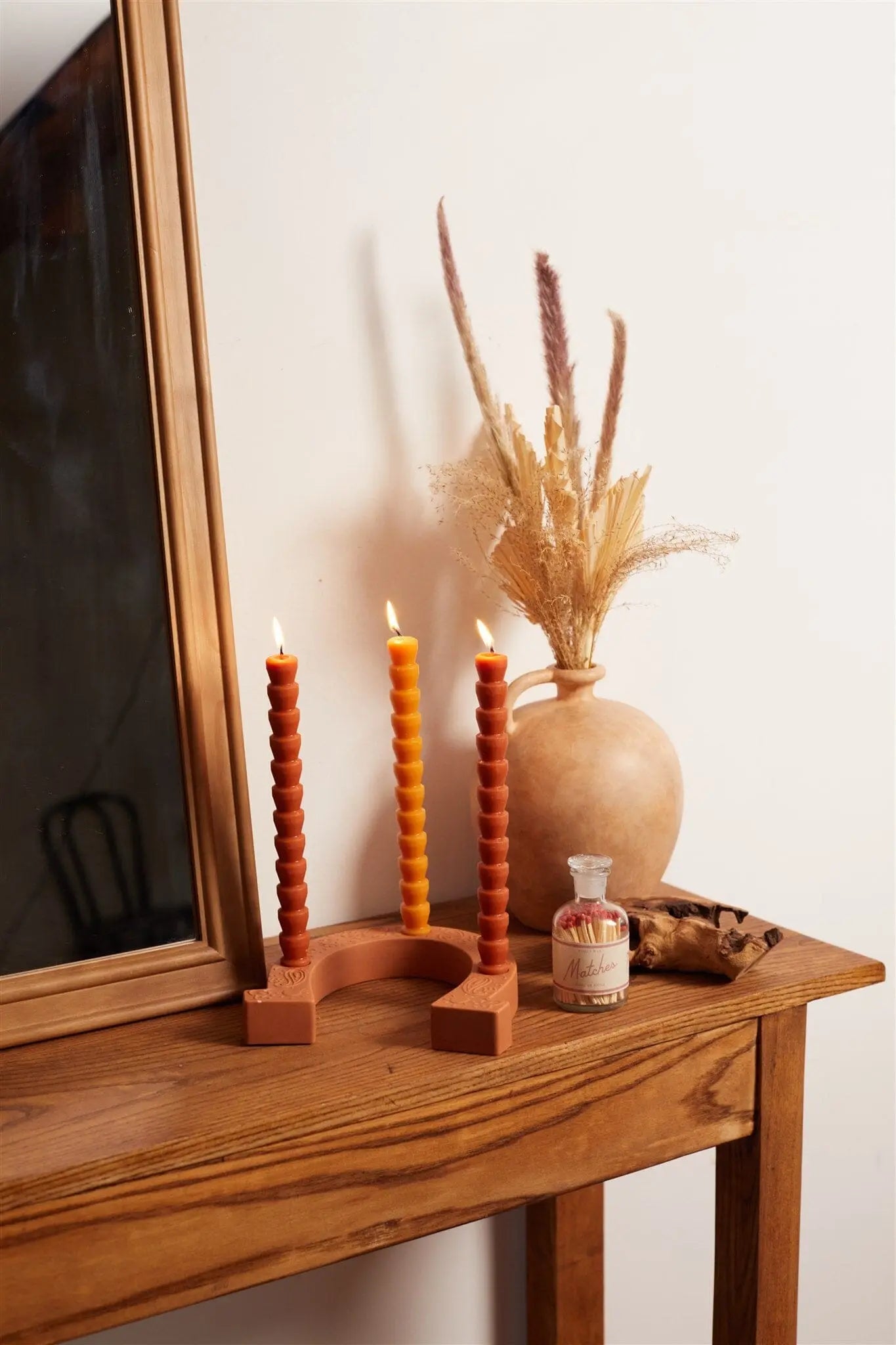 A wooden table holds three lit spirals in the Nashville Terracotta Ceramic Horseshoe Taper Holder, plus a bottle, dried grasses in a vase, and part of a framed mirror against a white wall for warm allure.