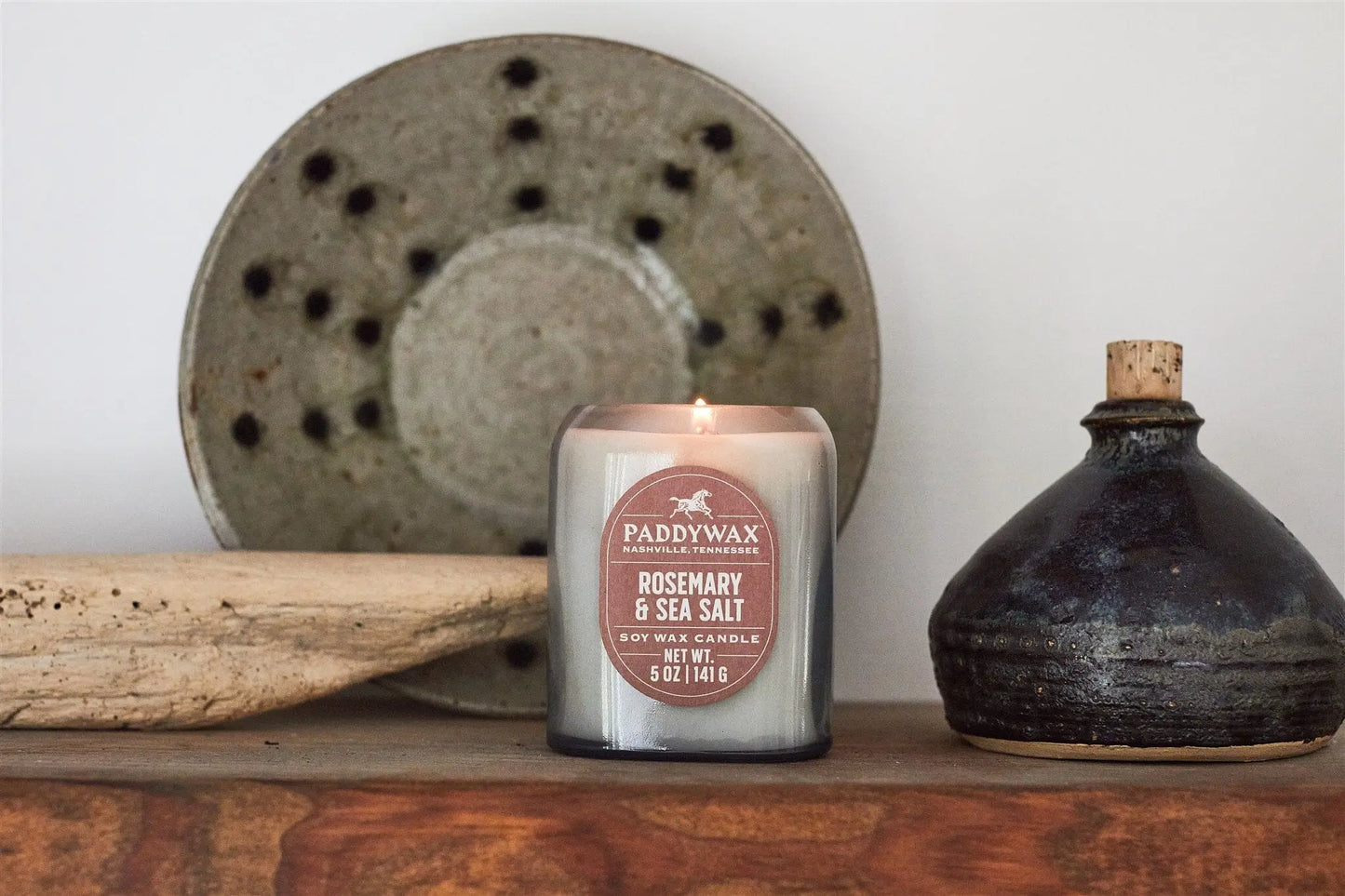 The Vista 5 oz Candle - Rosemary & Sea Salt sits on a wooden surface beside a ceramic plate, driftwood, and a dark ceramic bottle—bringing luxurious fragrance to your modern home.