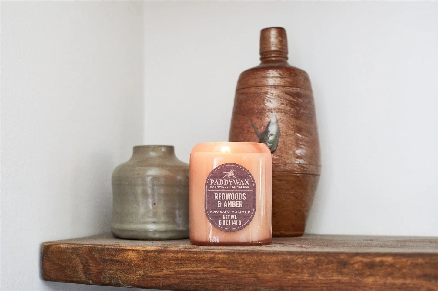 The Vista 5 oz Candle - Redwoods & Amber sits on a wooden shelf beside two ceramic vases against a white wall, making it an ideal addition to modern home decor.