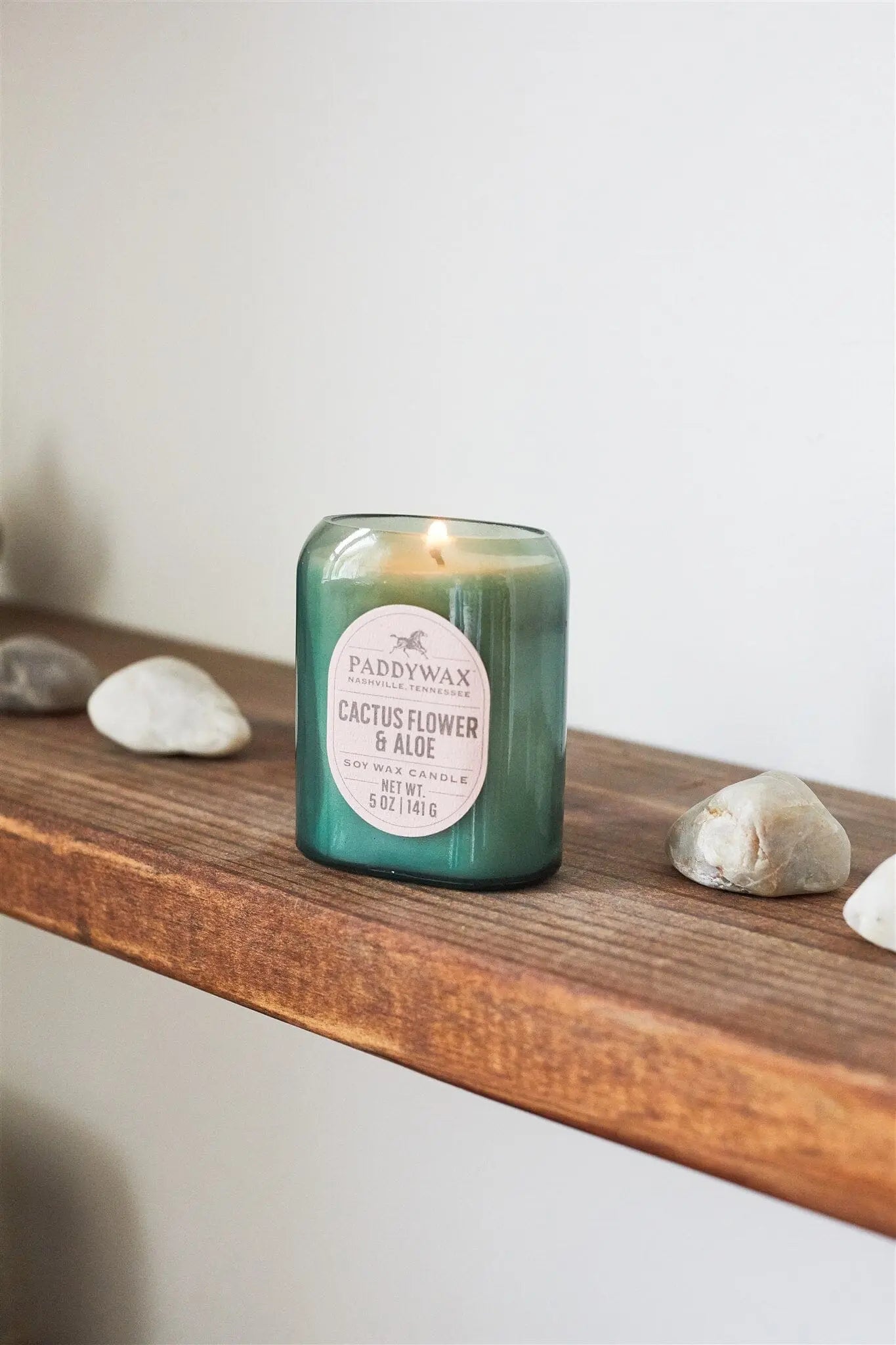 The Vista 5 oz Candle - Cactus Flower & Aloe is lit and placed on a wooden shelf with white and beige stones, bringing a luxurious fragrance and modern style to your home décor.