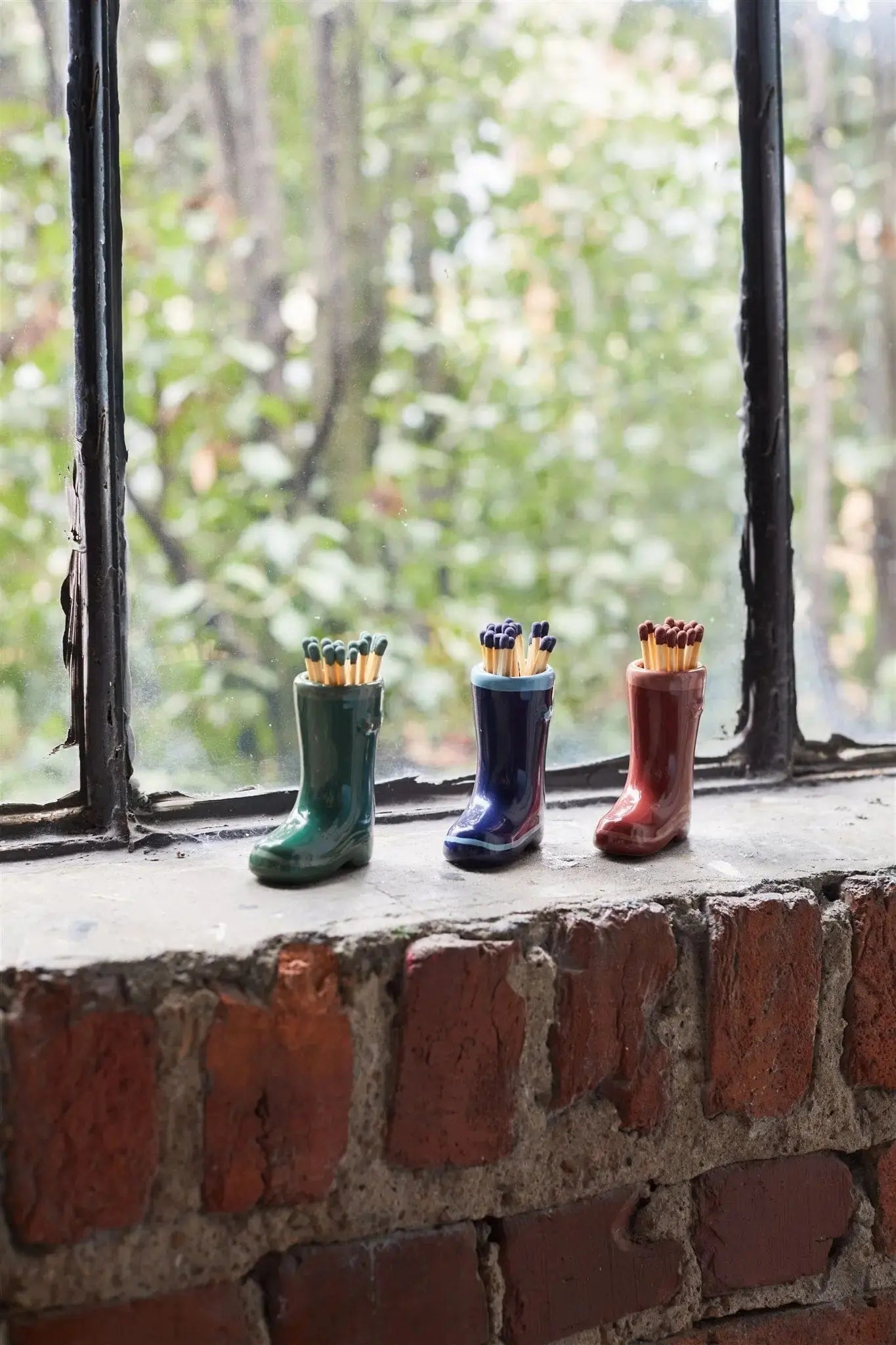 Three Wellington Boot Matches - Dark Green, each filled with safety matches, sit on a brick windowsill in front of a window with leafy trees outside.