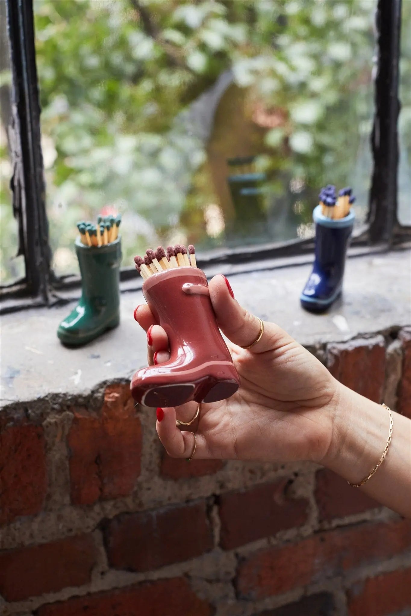 A hand holds the Wellington Boot Matches - Dark Red match holder filled with matches, while green and blue boot-shaped holders sit on a windowsill, creating a delightful home decor gift.
