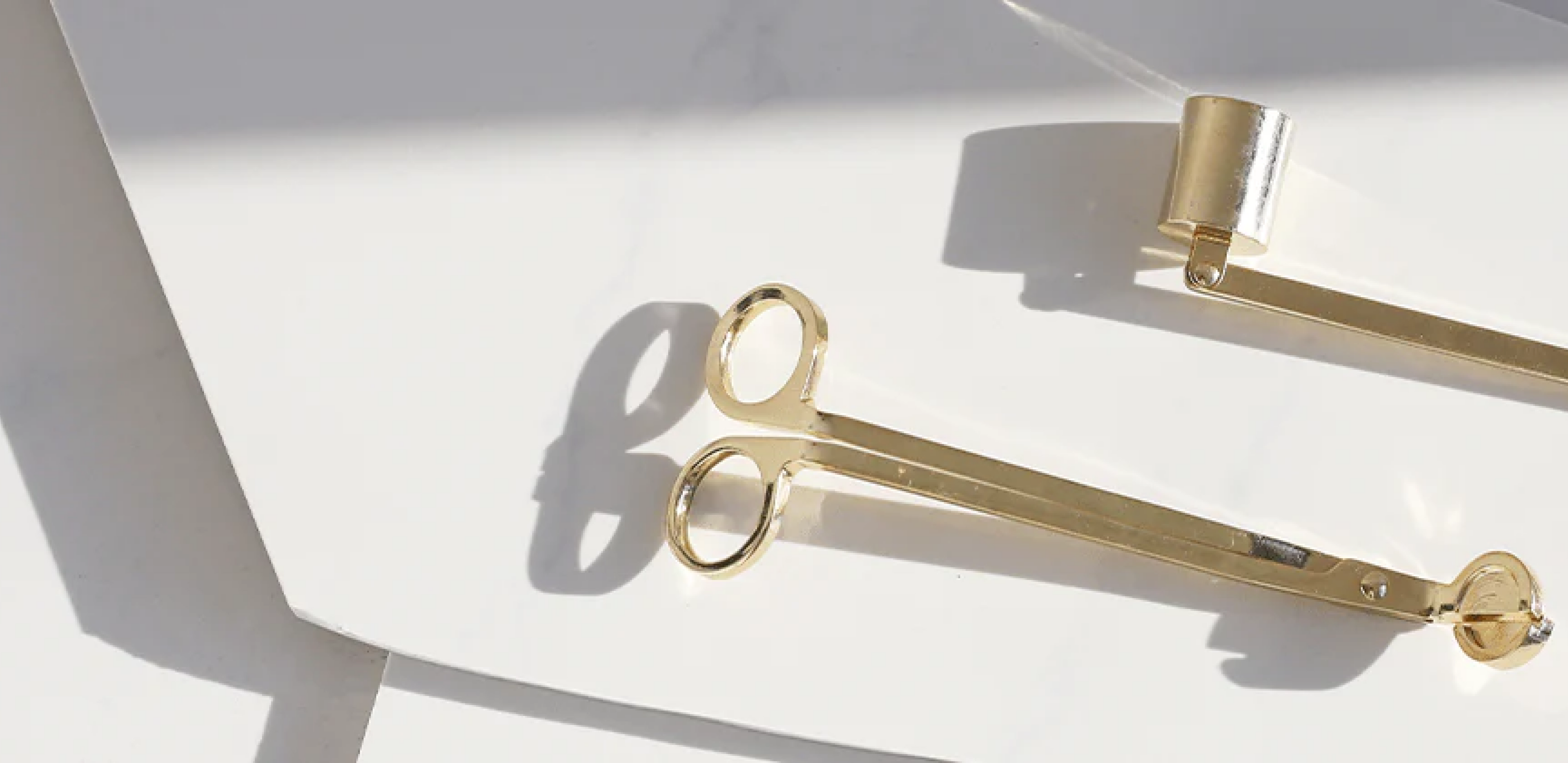 A gold candle wick trimmer and snuffer rest on white sheets, illuminated by soft natural light.