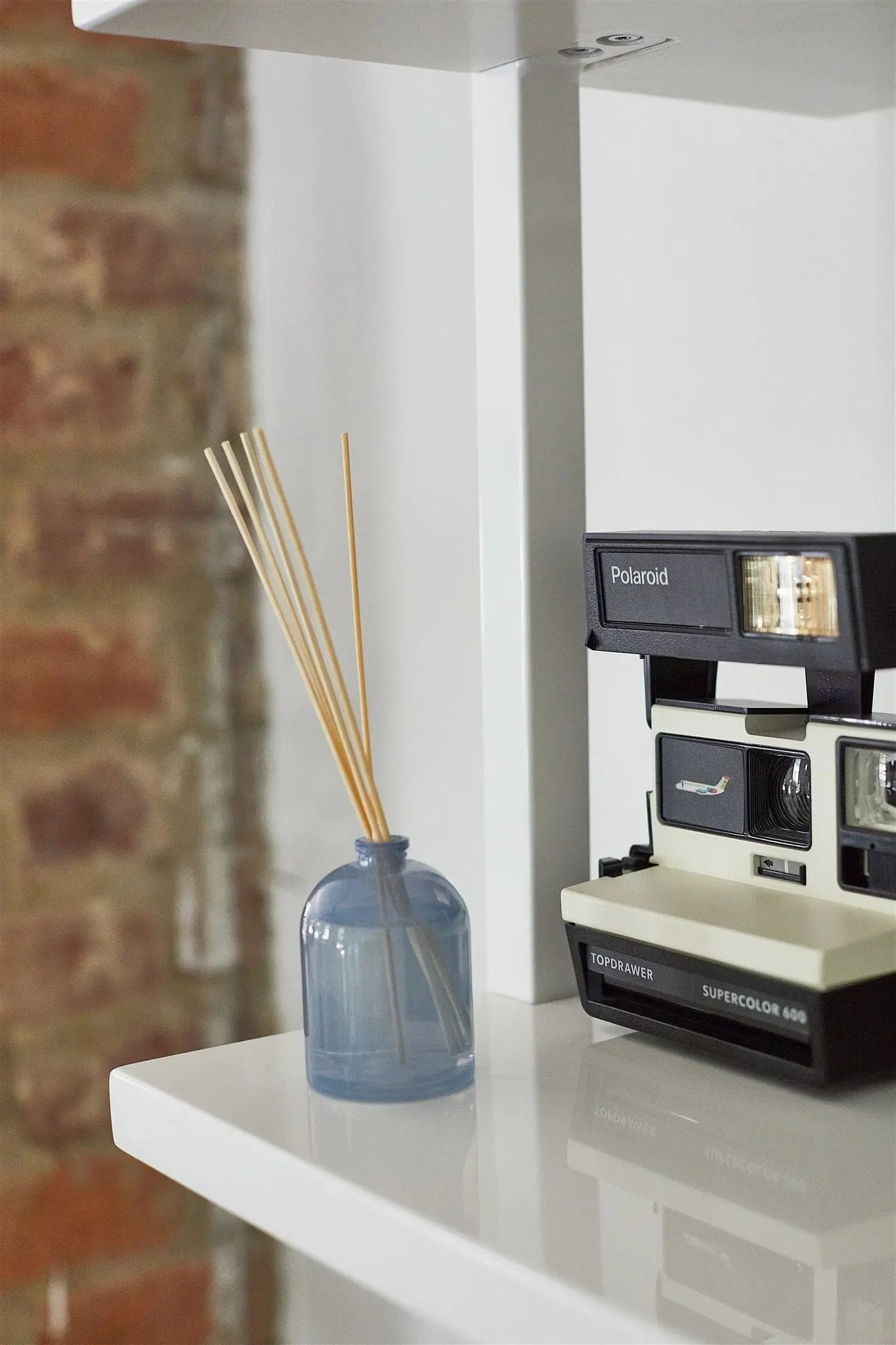 A Petite Reed Diffuser - Wild Fig with blue glass and fragrance reeds sits on a white shelf beside a vintage Polaroid Supercolor 645CL instant camera, all set against a brick wall backdrop.