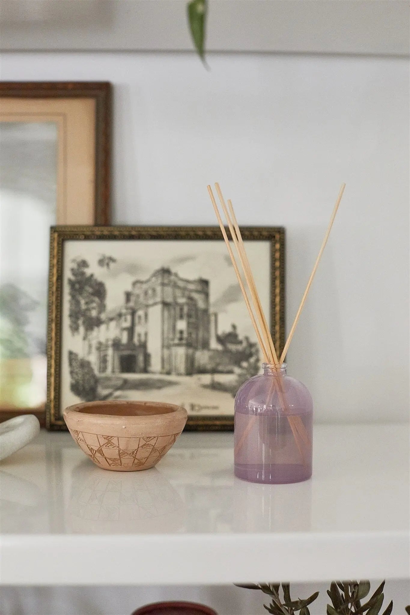 A small ceramic bowl and the Petite Reed Diffuser - Lavender sit on a white surface before a framed black-and-white architectural drawing, creating a stylish, giftable accent for any space.