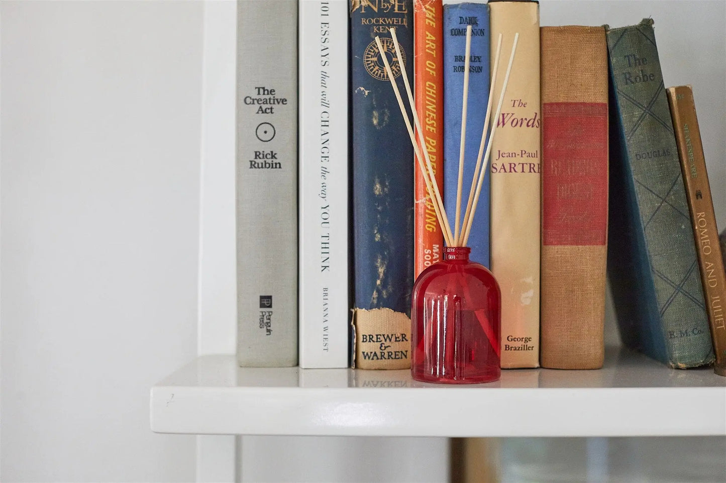 The Petite Reed Diffuser - Cranberry sits on a white shelf in front of vertically stacked colorful books, filling the air with its delightful fragrance.
