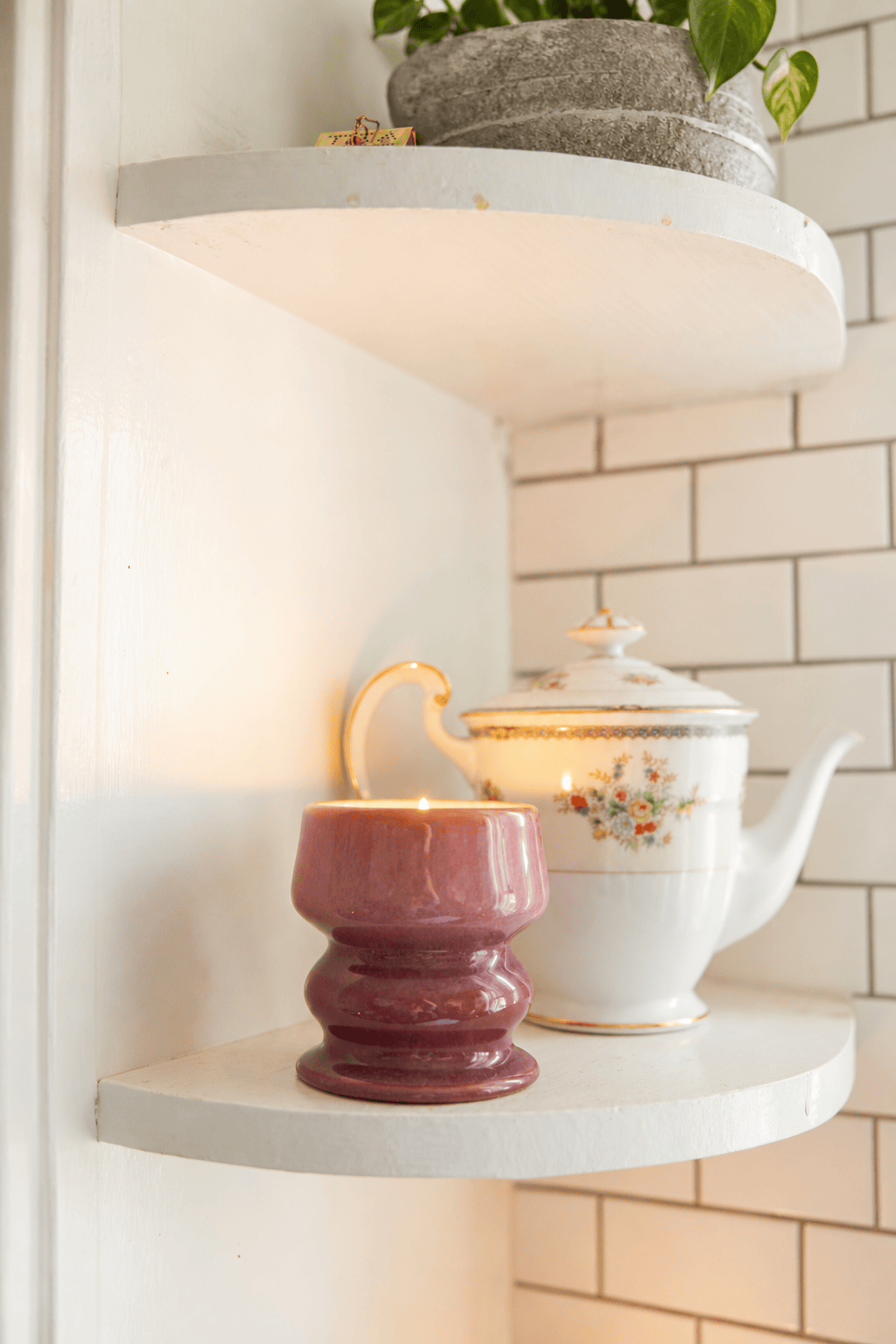 A lit Wild Fig & Cedar - Melt 8.5 oz Candle in a ceramic holder sits on a white shelf next to a floral-patterned teapot with a gold handle. A small green plant in a gray pot rests on the shelf above, set against a white tiled wall in the background.