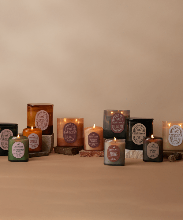 A selection of lit and unlit candles in various colored jars and sizes are arranged on pieces of stone and brick against a neutral beige background.