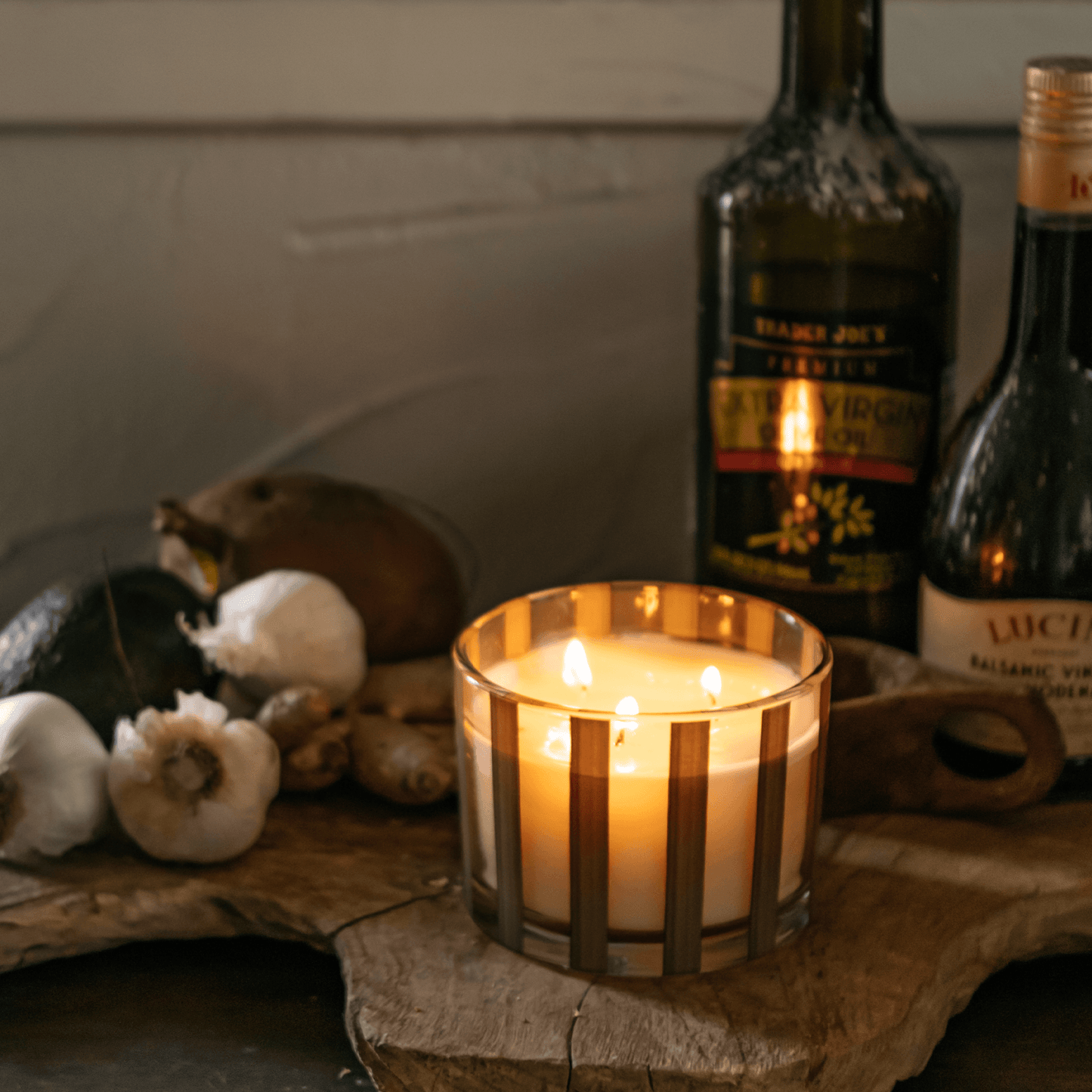 Striped brown clear glass vessel with white wax and three wicks; a part of the Al Fresco collection; comes with woven rattan lid; pictured lit on a cutting board full of Mediterranean ingredients..