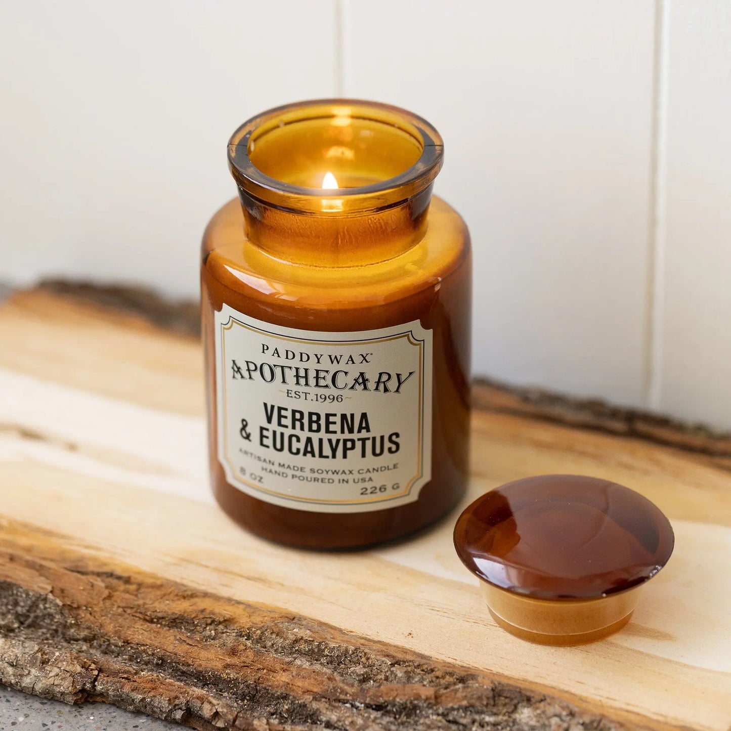 8 oz brown apothecary bottle glass vessel with a vintage-style label; white wax; part of the apothecary collection; candle lid off and scented candle burning on wood piece