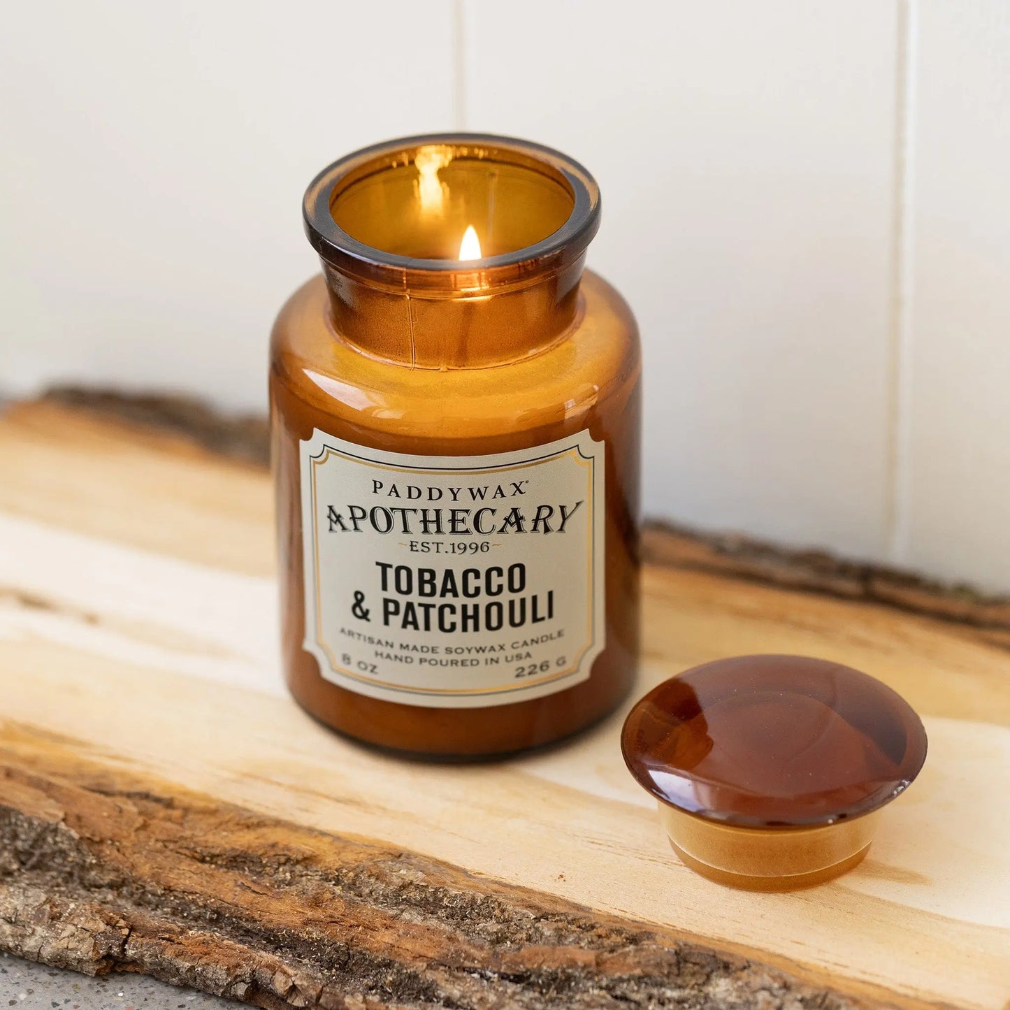 8 oz brown apothecary bottle glass vessel with a vintage-style label; white wax; part of the apothecary collection; candle lid off and scented candle burning on wood piece