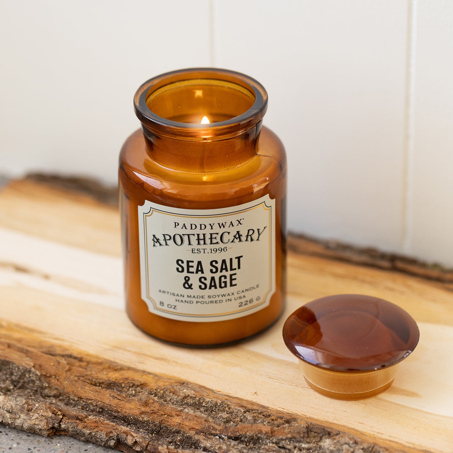 8 oz brown apothecary bottle glass vessel with a vintage-style label; white wax; part of the apothecary collection; candle lid off and scented candle burning on wood piece