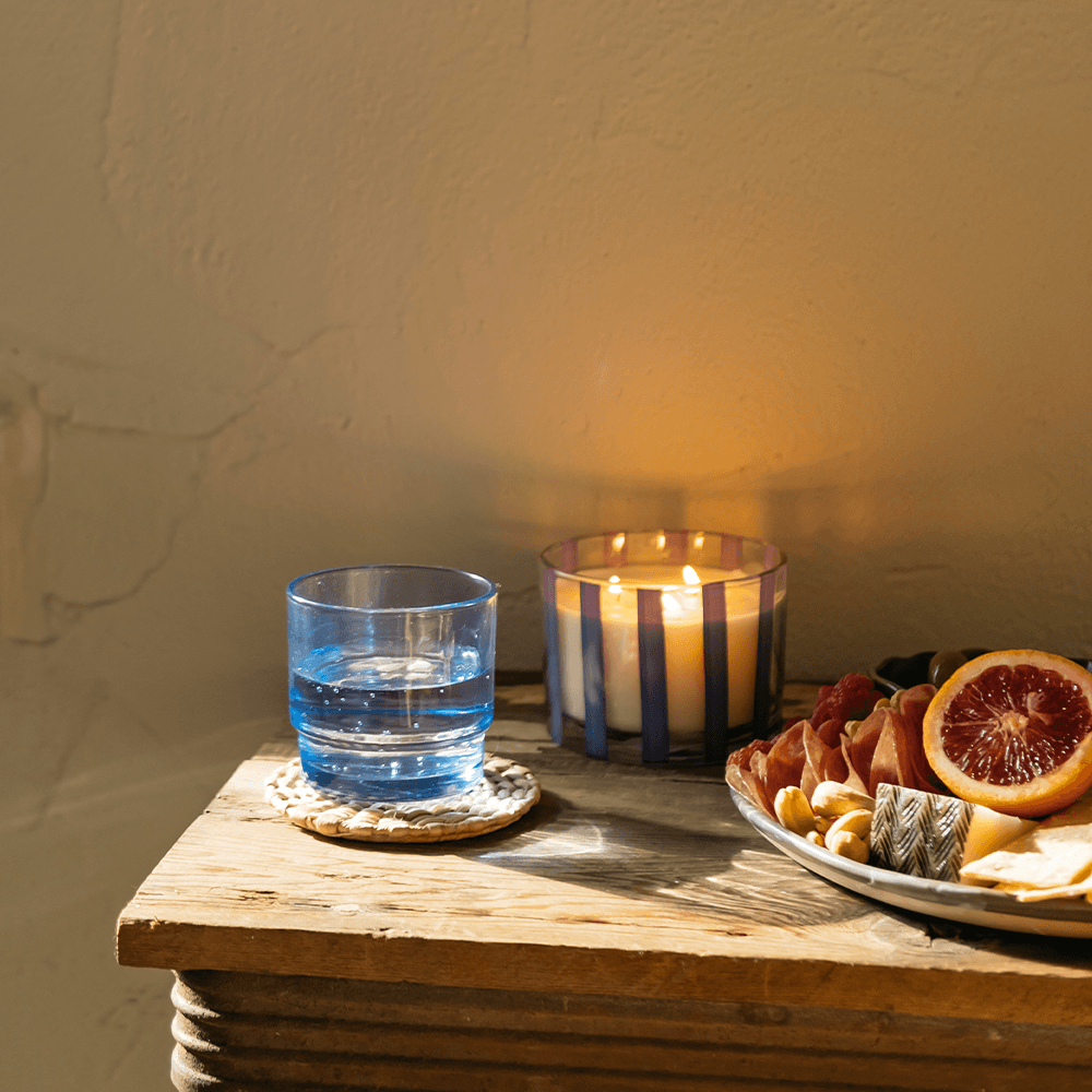 12 oz clear glass vessel with pink/purple stripes, white wax, and three wicks; a part of the Al Fresco collection; comes with woven rattan lid; pictured lit next to a meal