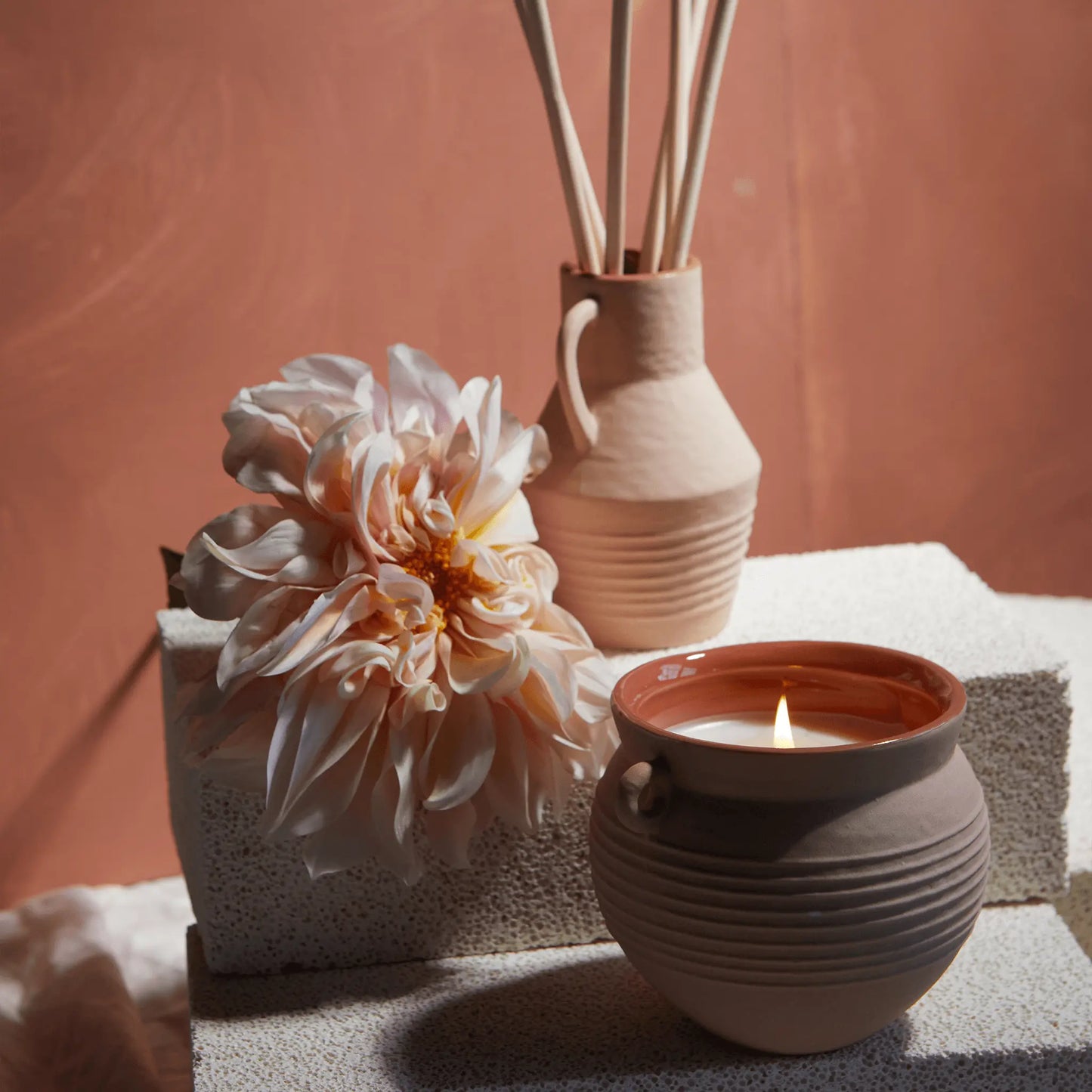 The Santorini 8.5 oz. Candle - Black Fig & Olive is displayed on textured white blocks against a muted pink background, alongside a ceramic jug with reed sticks and a large pale flower from the Santorini collection.