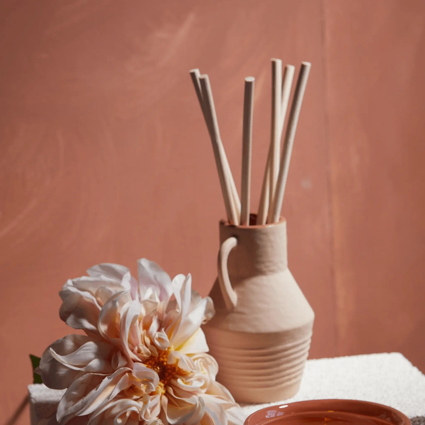 The Santorini 4 fl oz. Diffuser - Black Fig & Olive is displayed with wooden sticks, a large pale flower, and a terracotta dish on a white textured surface against a muted pink background.