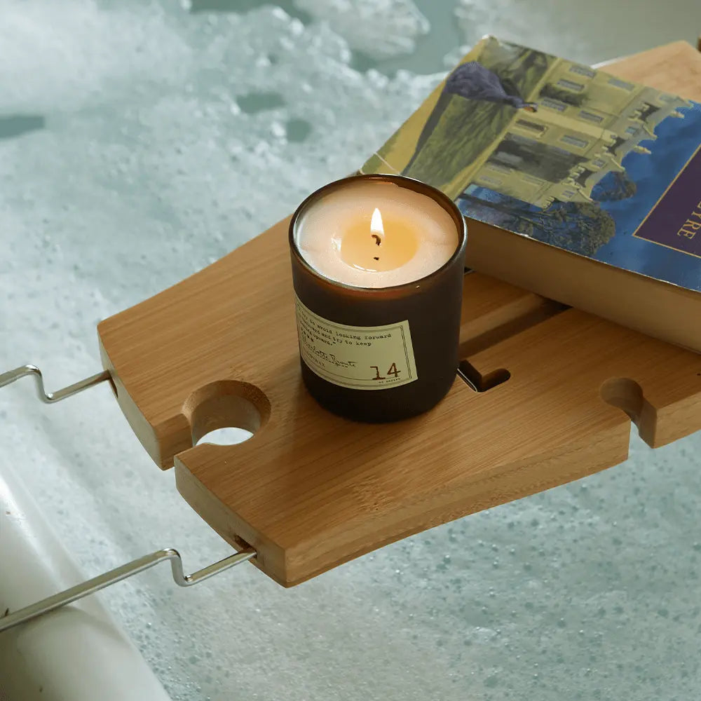 A Library 6 oz Candle – Charlotte Bronte and a book rest on a wooden bathtub tray above foamy bathwater, filling the air with exquisite fragrance.