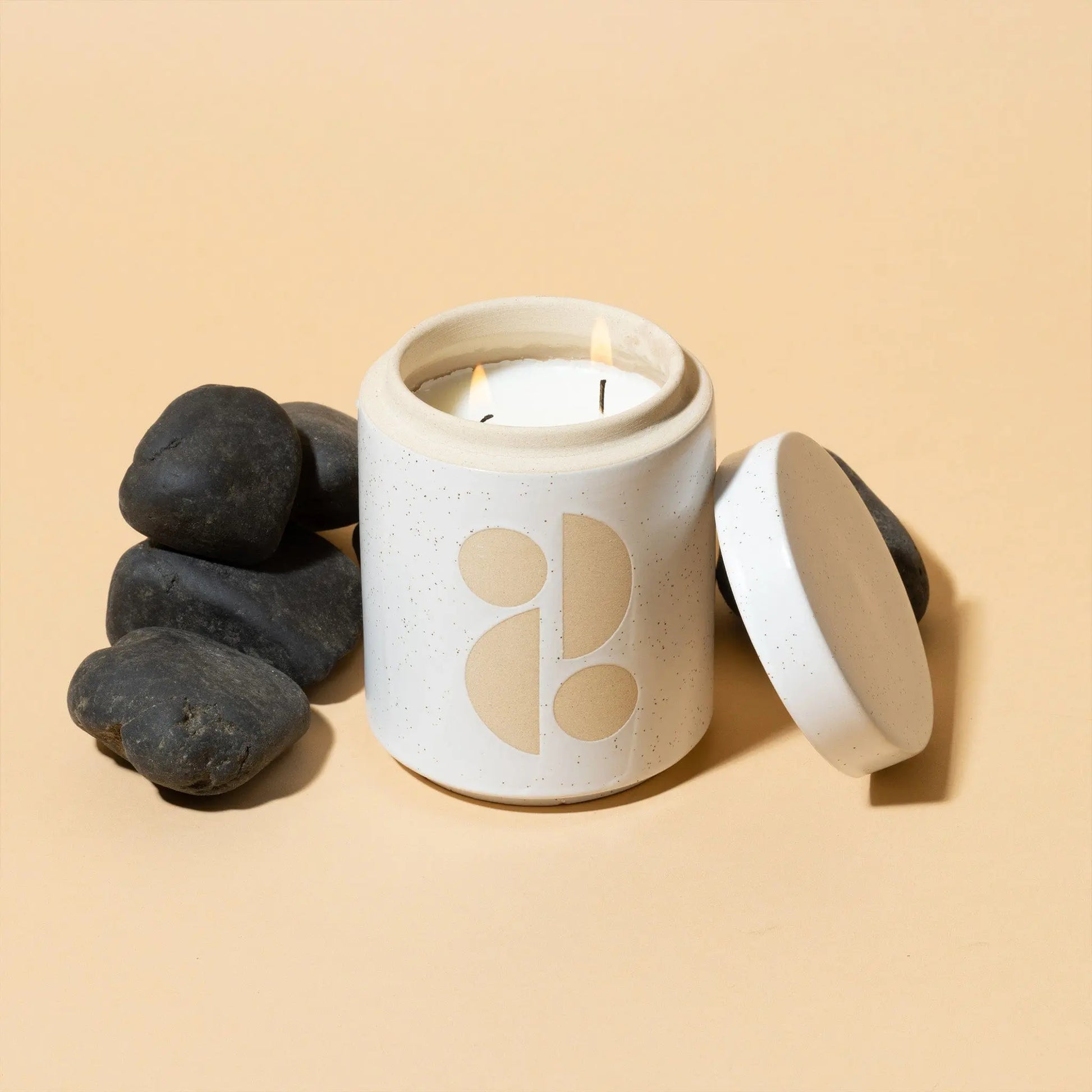 The Form 12 oz Candle - Tobacco Flower, featuring a white ceramic minimalist design and matching lid, rests beside smooth black stones on a beige background—an elegant upgrade for your home fragrance collection.