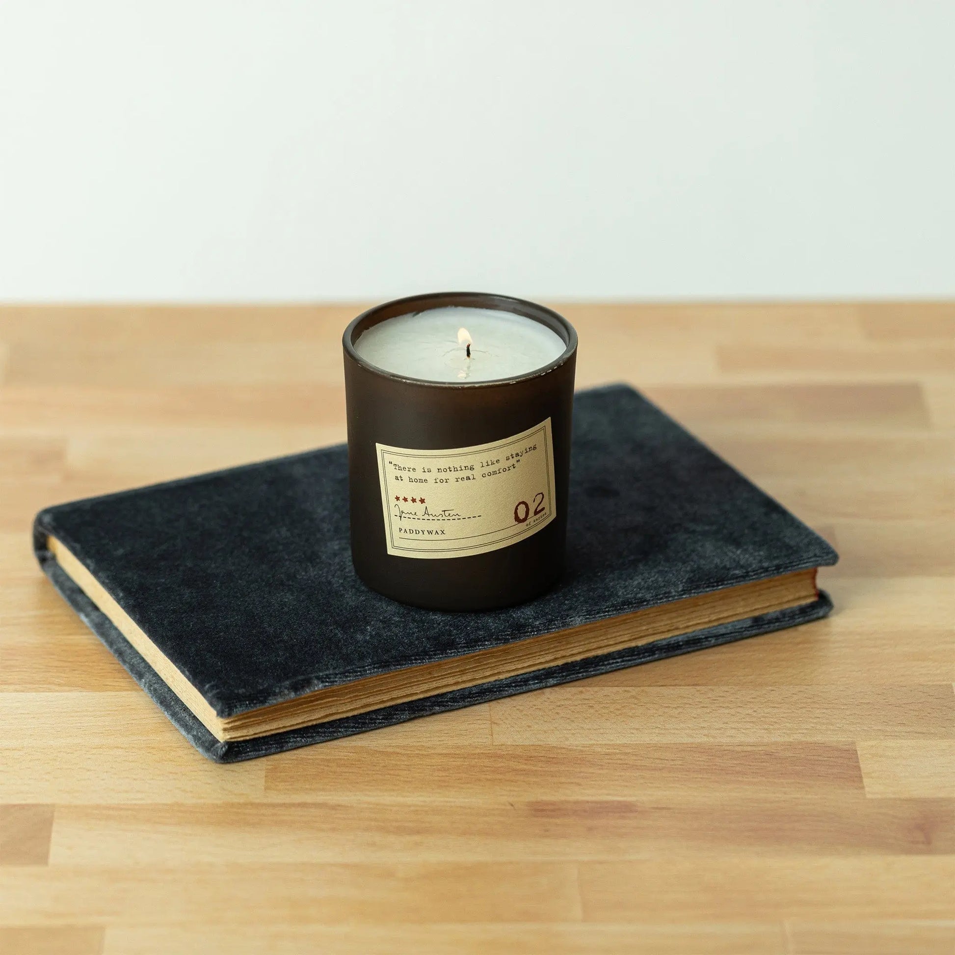 The Library 6 oz Candle – Jane Austen envelops the air with bookish scents as it rests on a closed, dark book—creating a warm scene for bibliophiles atop a wooden surface.