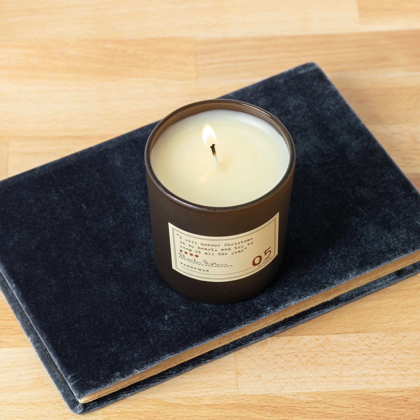 The Library 6 oz Candle – Charles Dickens sits in a dark glass jar on a closed dark gray velvet book, offering literary fragrance for book lovers atop a light wooden surface.
