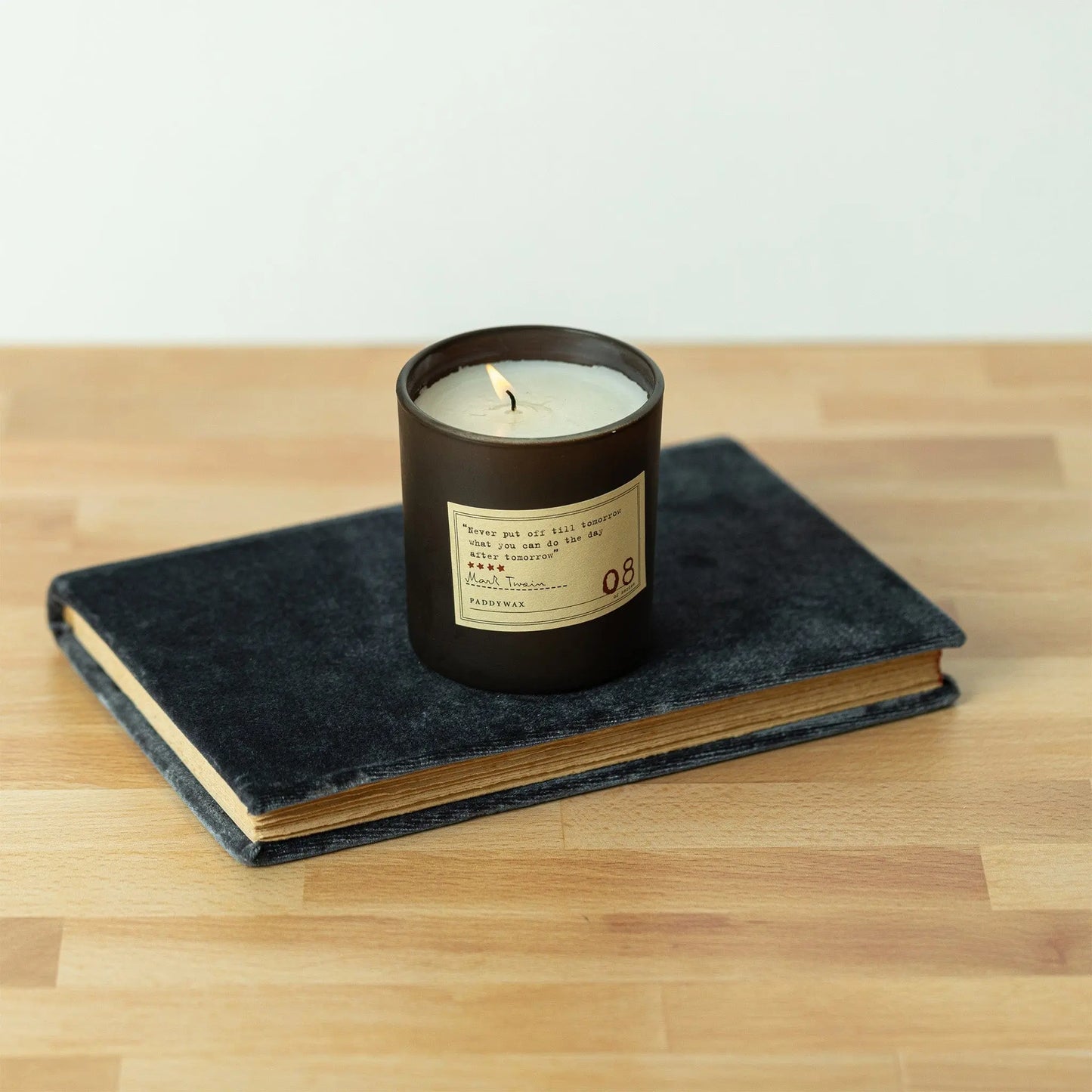 The Library 6 oz Candle - Mark Twain, in a dark glass jar with rich fragrance, rests atop a closed black book on a wooden surface.