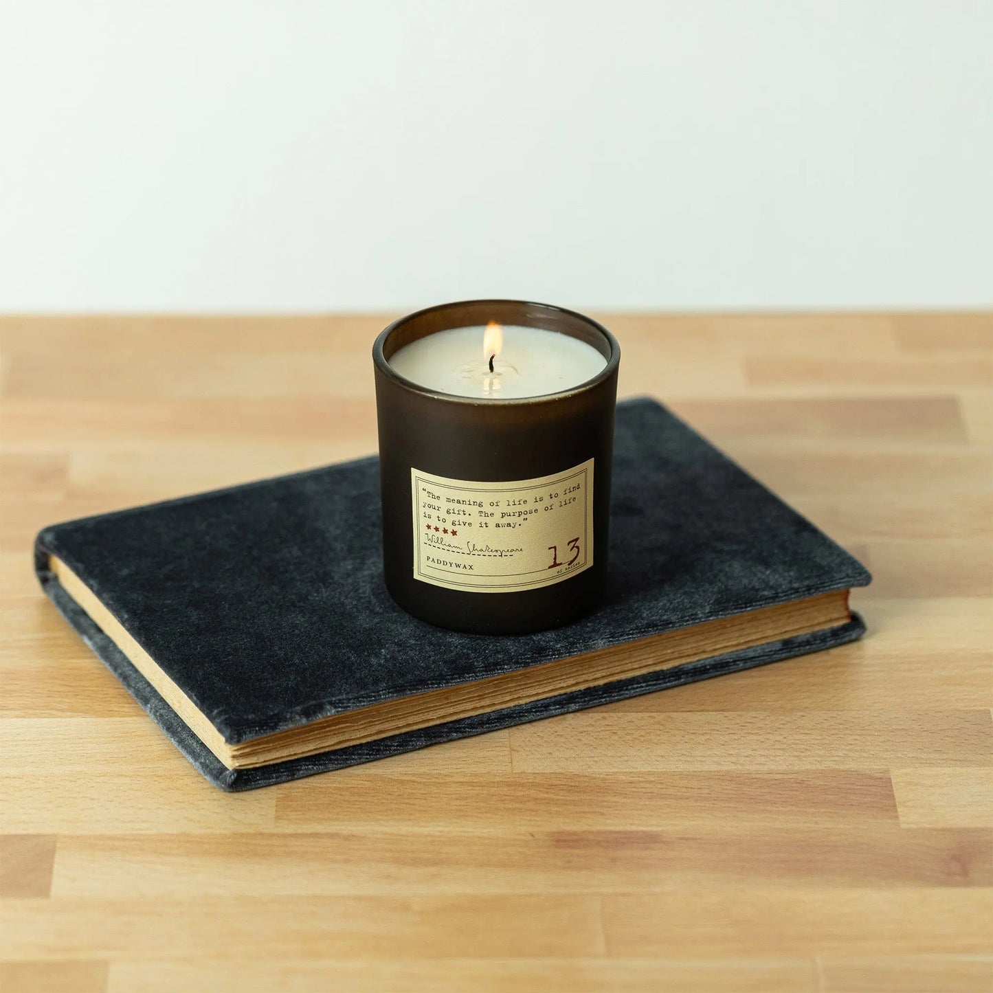 A Library 6 oz Candle - William Shakespeare, renowned for its exquisite fragrance, sits in a dark glass container atop a closed hardcover book on a wooden surface.