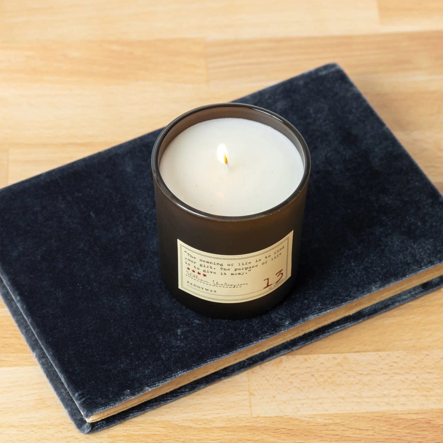The Library 6 oz Candle - William Shakespeare, in a dark glass holder, rests unlit atop a closed, textured dark book on a light wooden surface.