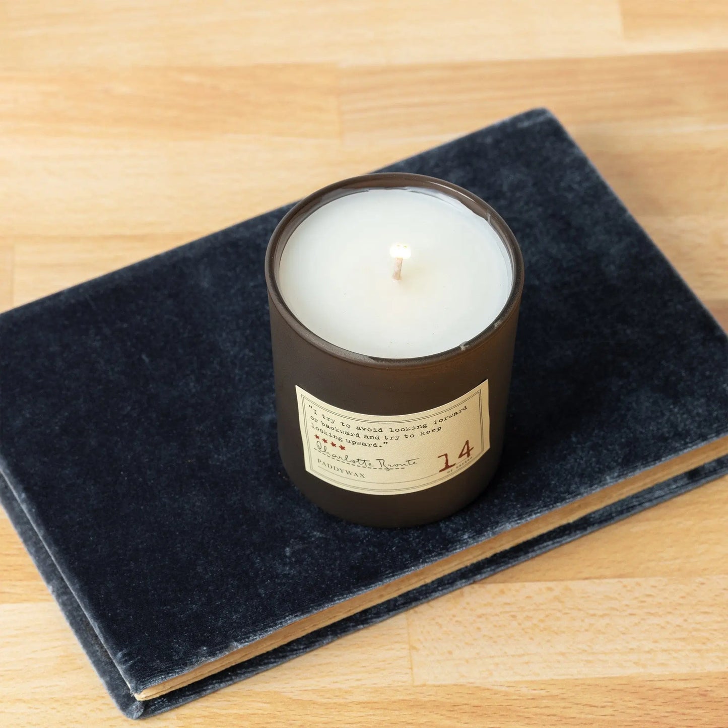 The Library 6 oz Candle - Charlotte Bronte, in a dark glass container, sits on a closed dark gray book atop a light wooden surface, filling the air with exquisite literary-inspired fragrance.