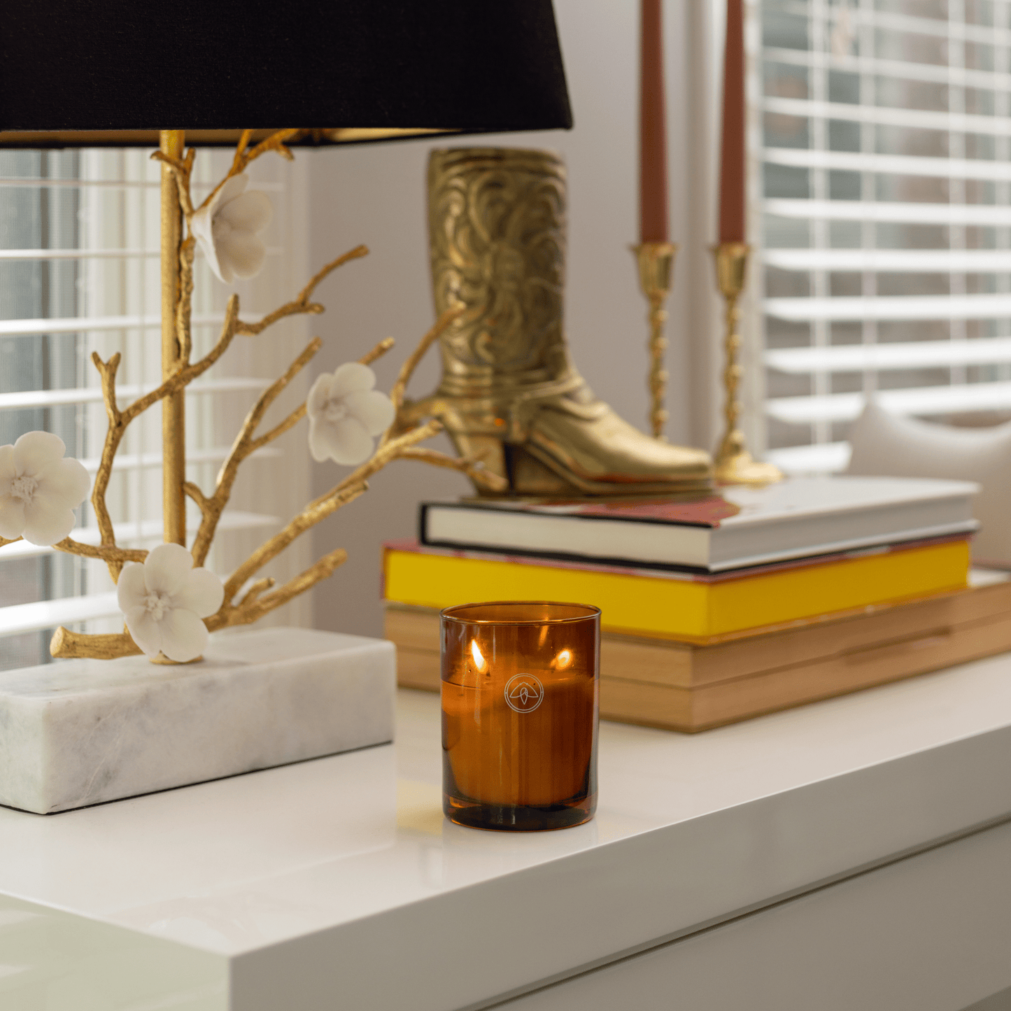 10oz Glow two wick candle on white table with lamp, books, metal cowboy boot decor and taper candlesticks all in front of a window