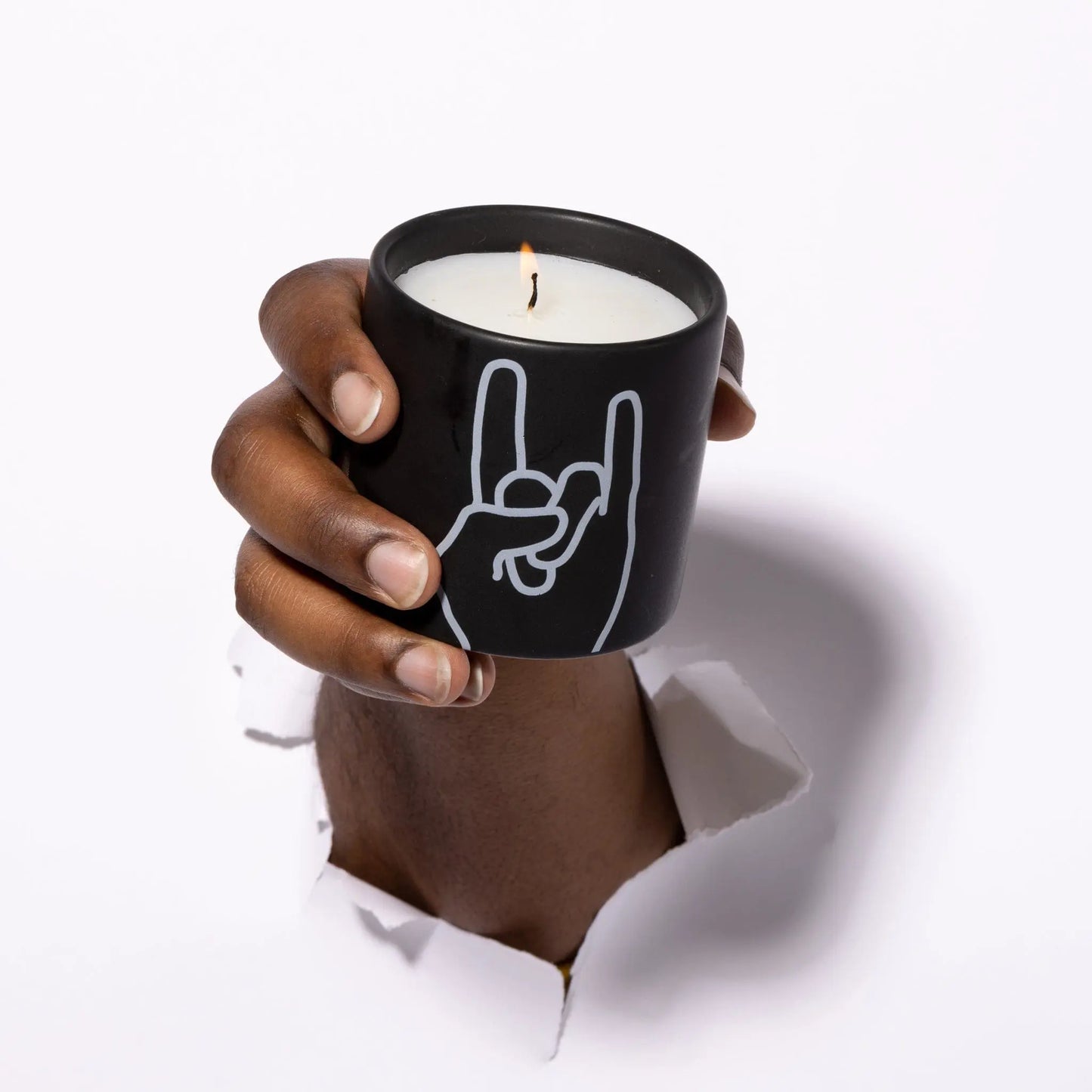 A hand emerges through a white surface, holding the Impressions 5.75 oz Candle - Leather + Oakmoss Rock On in a matte ceramic jar, decorated with a white line art of the rock on gesture.